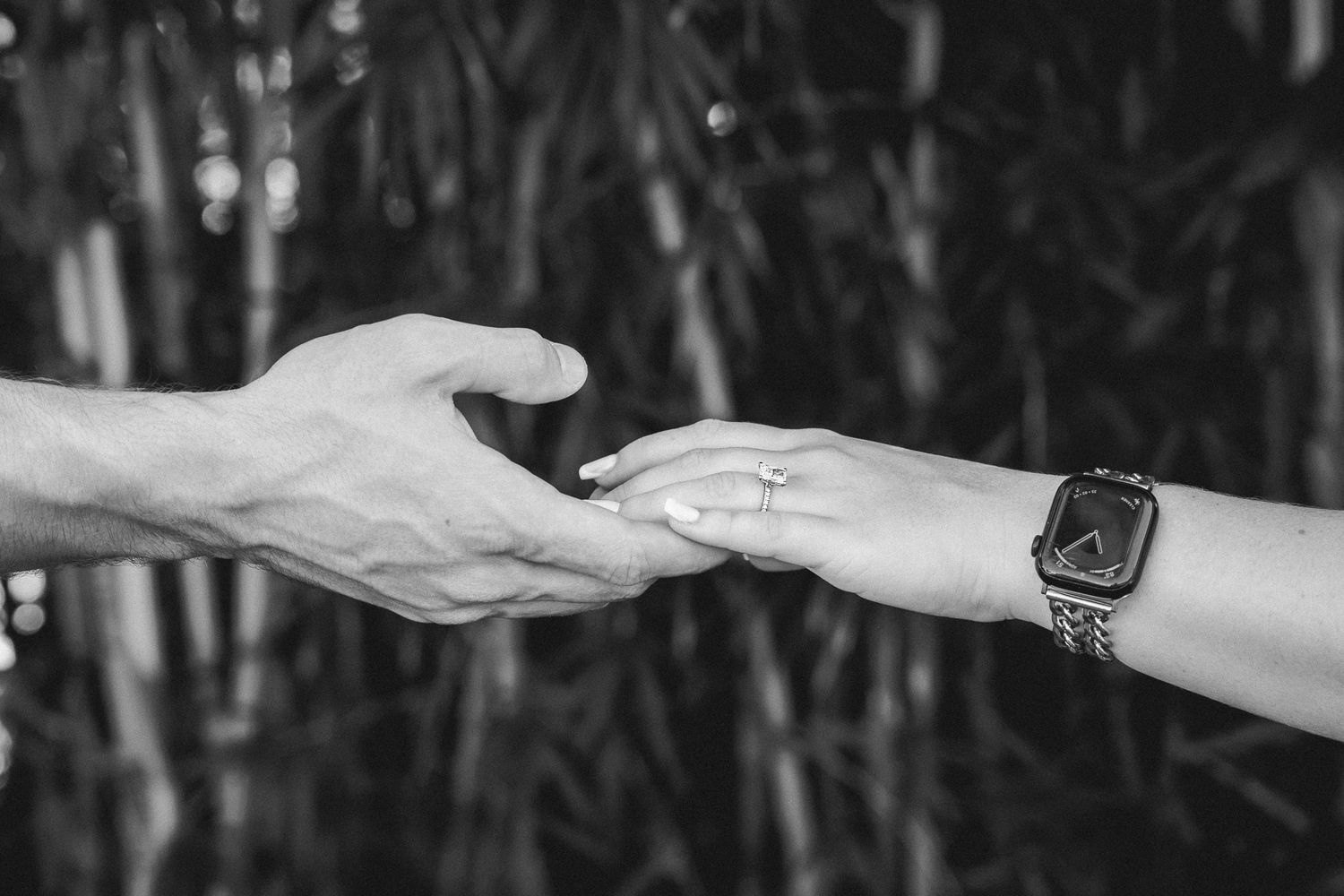 Close-up black and white photo of a couple holding hands, showcasing an engagement ring and a smartwatch.
