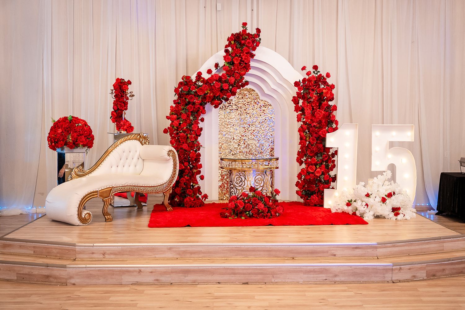 Elegant quinceañera stage with red rose arch, white chaise lounge, gold birdcage, and decorative number 15 on wooden platform.