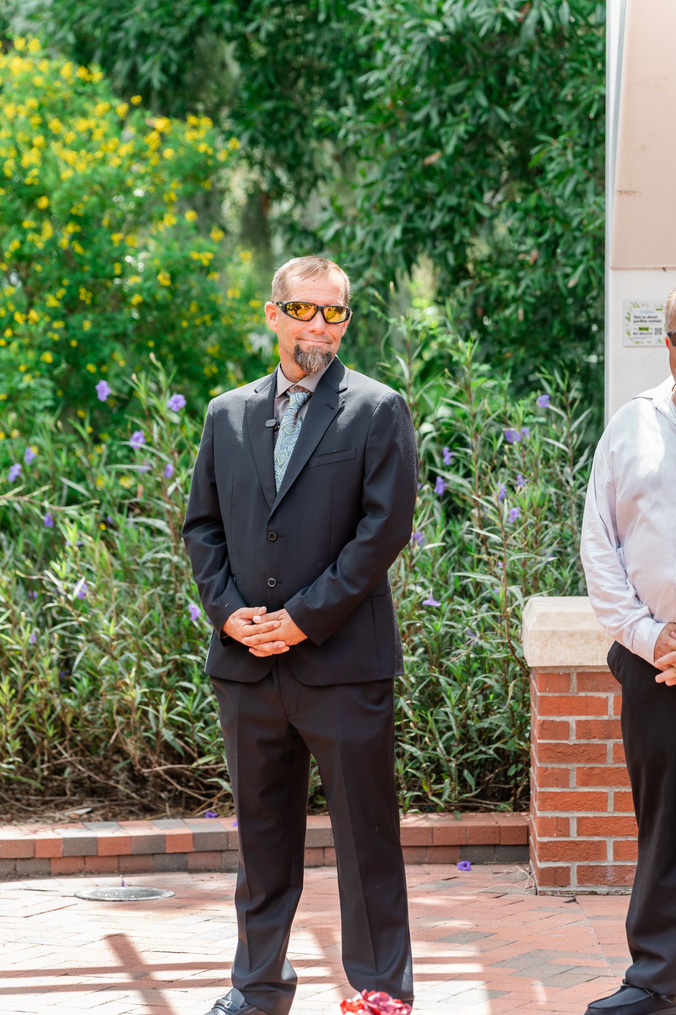 Man in black suit with facial tattoos and orange sunglasses standing outdoors near brick pillar and lush green garden.