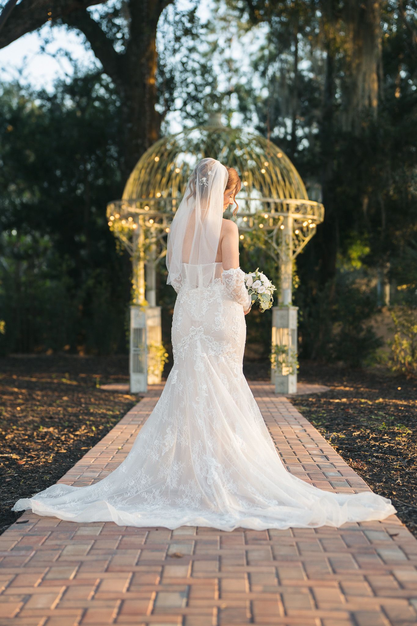 Bride in lace mermaid gown with long veil stands before illuminated garden arch at dusk.