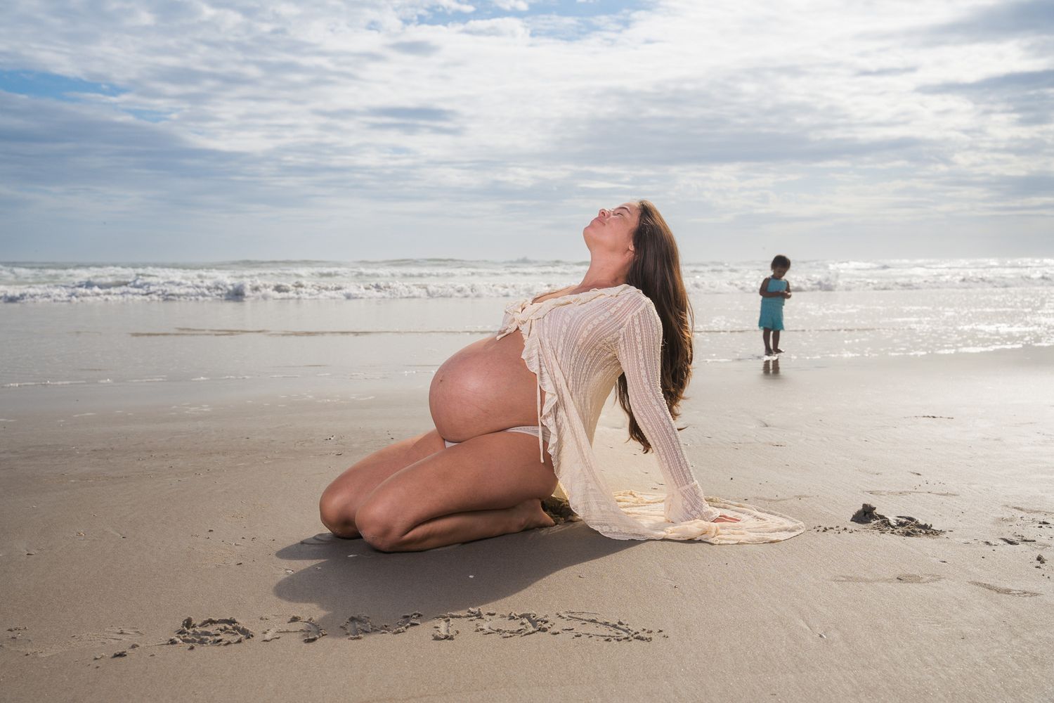 Pregnant woman kneels on beach in white lace cover-up while child plays near ocean waves.