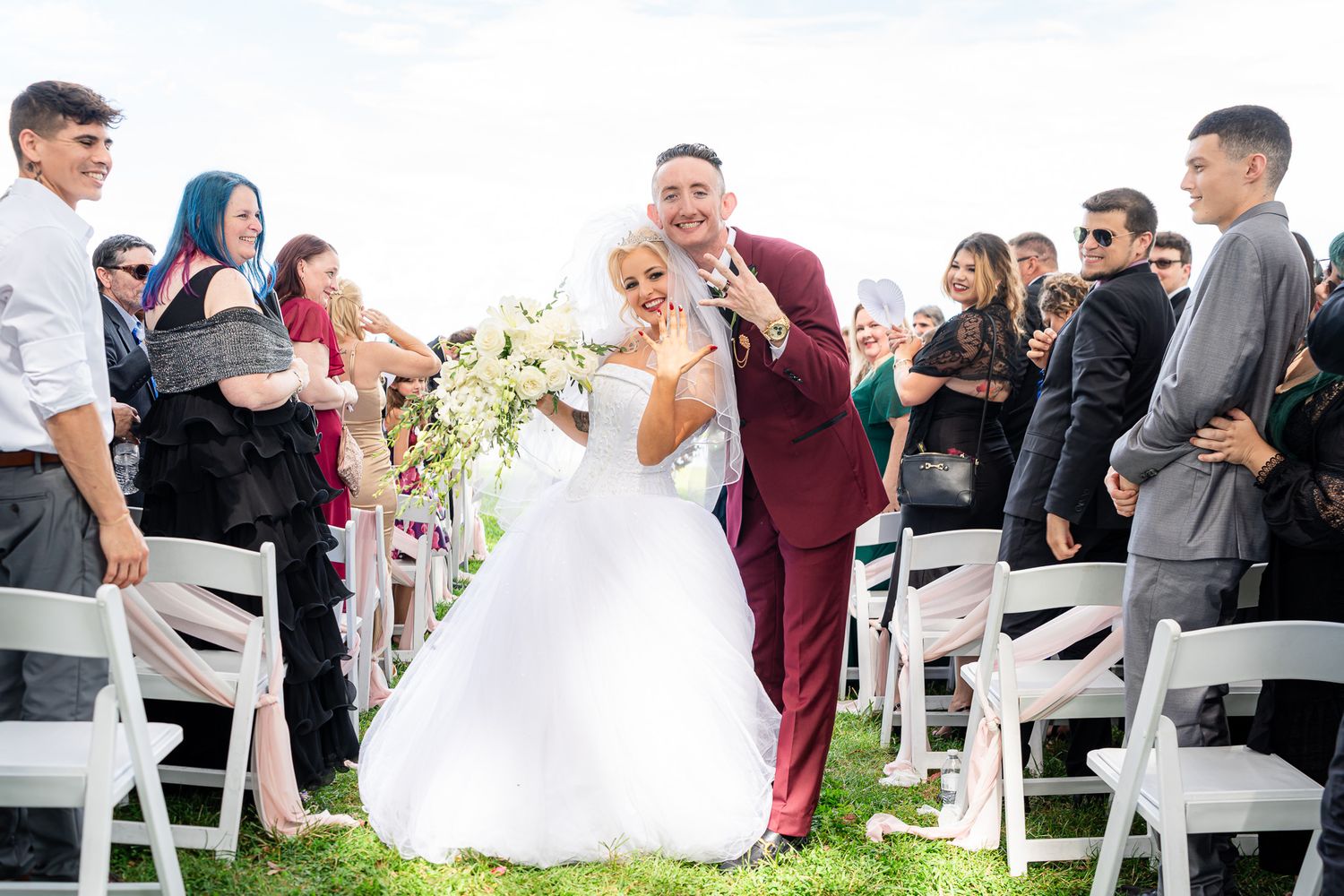 Newlyweds walk joyfully down the aisle as guests cheer at their outdoor lakeside wedding ceremony.
