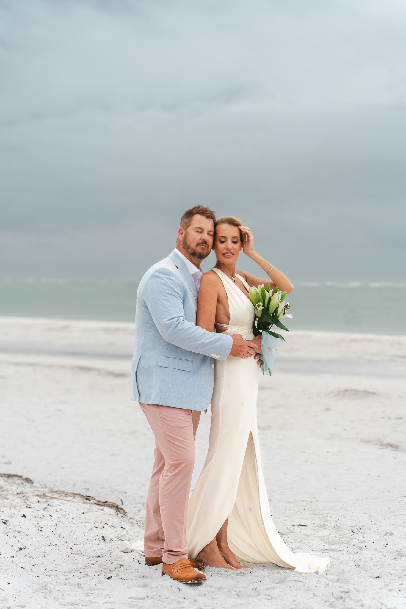 Bride and groom embracing on a white sand beach, bride holding a bouquet, groom in light blue suit and pink pants.