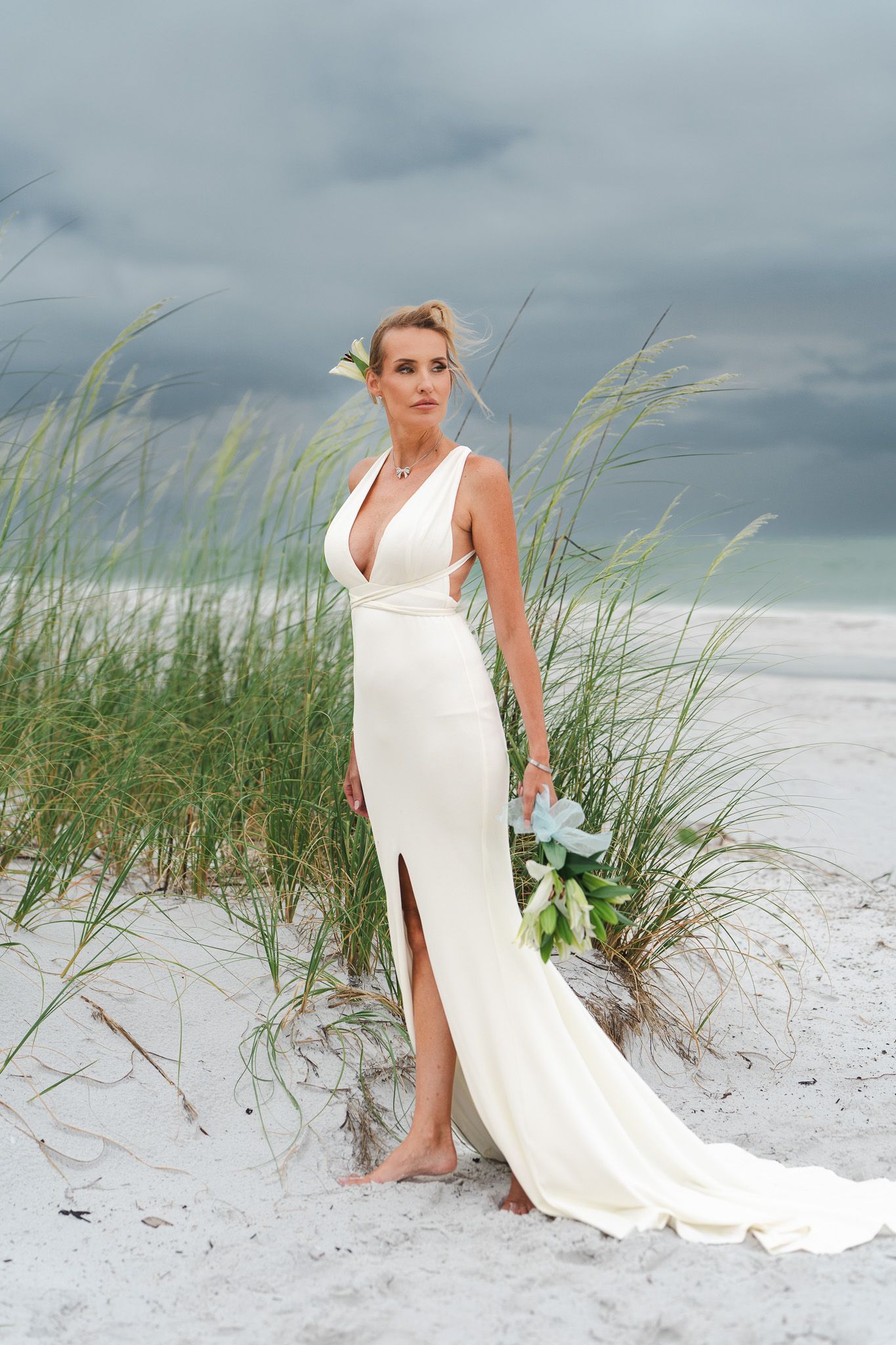 Bride in elegant white deep-V wedding gown holding bouquet on sandy beach with sea grass and stormy sky.