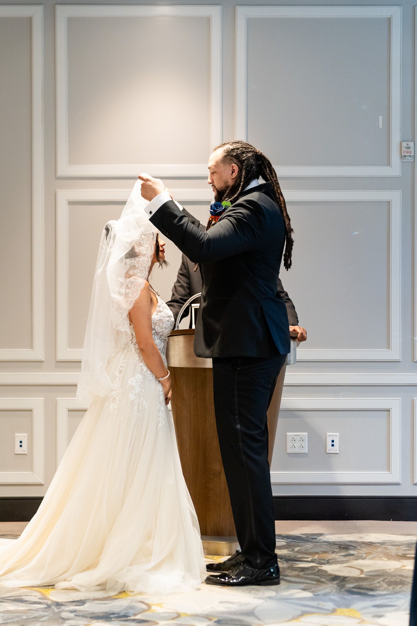 Groom with dreadlocks lifts brides veil tenderly during intimate indoor wedding ceremony moment.