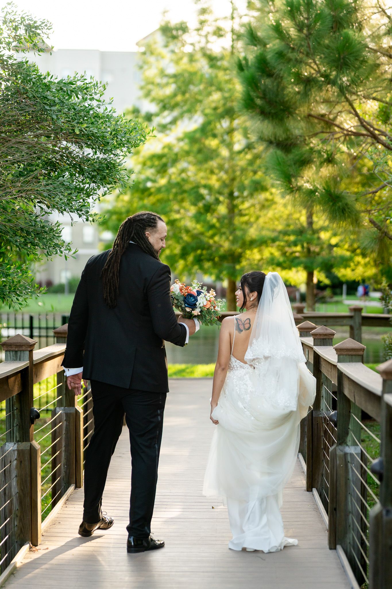Bride and groom walk hand-in-hand on wooden bridge surrounded by lush green trees.