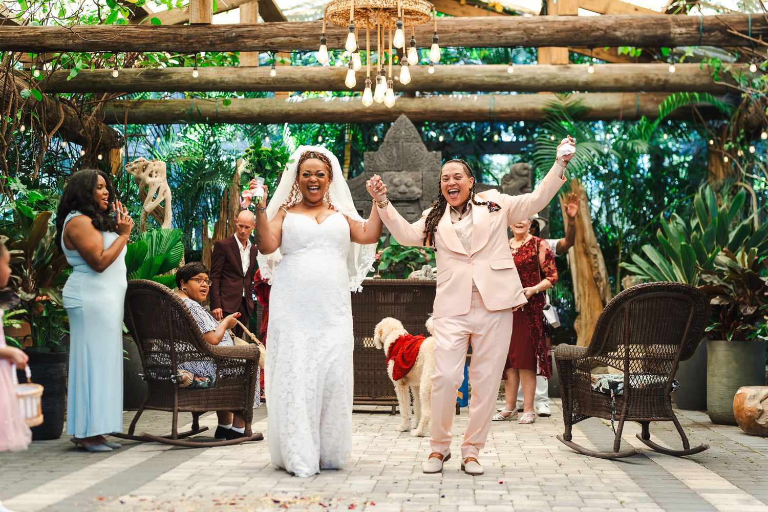 Two brides celebrate joyfully at outdoor garden wedding ceremony under rustic pergola with lush greenery.
