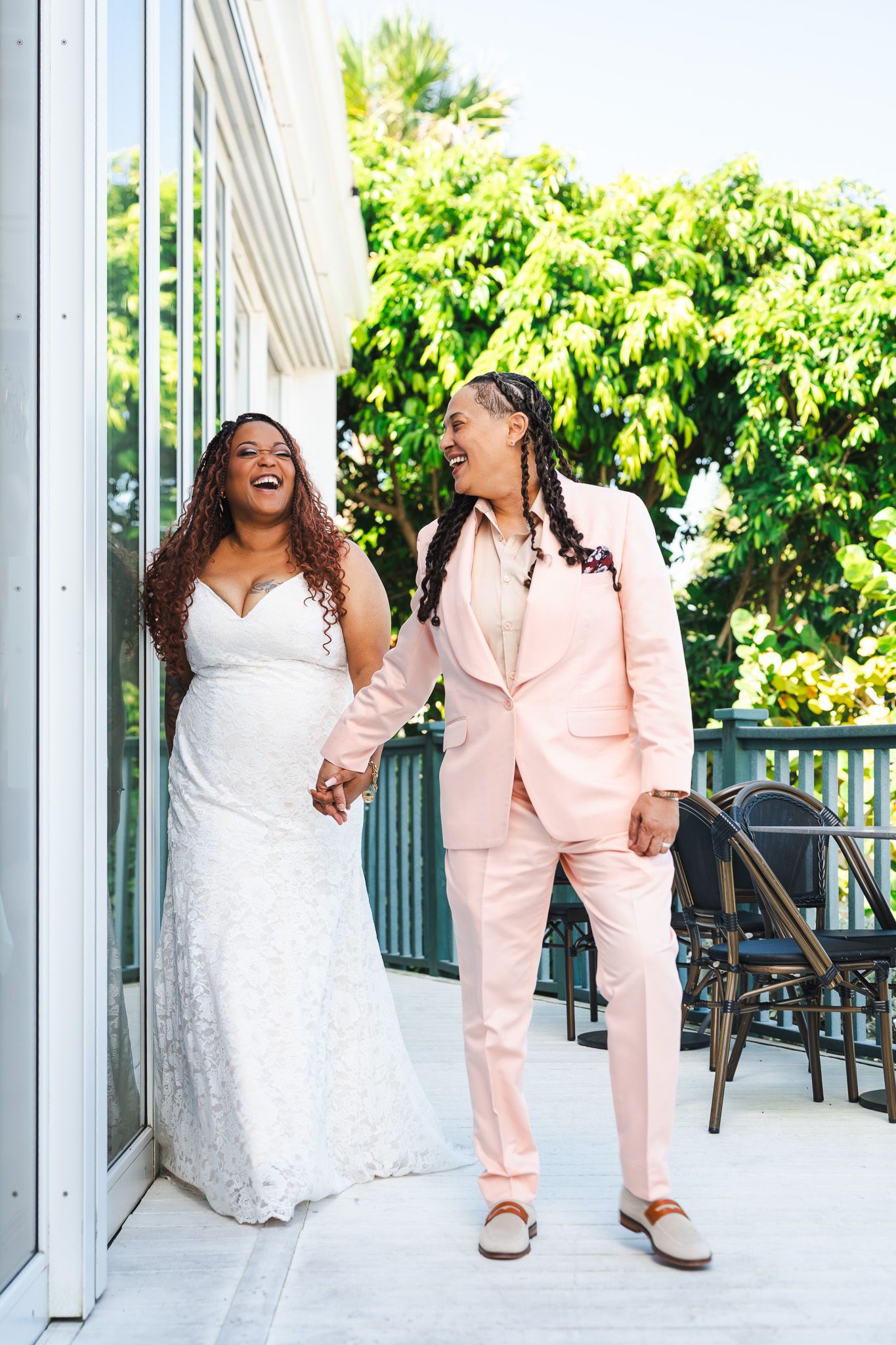 Two brides laugh and hold hands walking on sunny outdoor terrace in wedding attire.