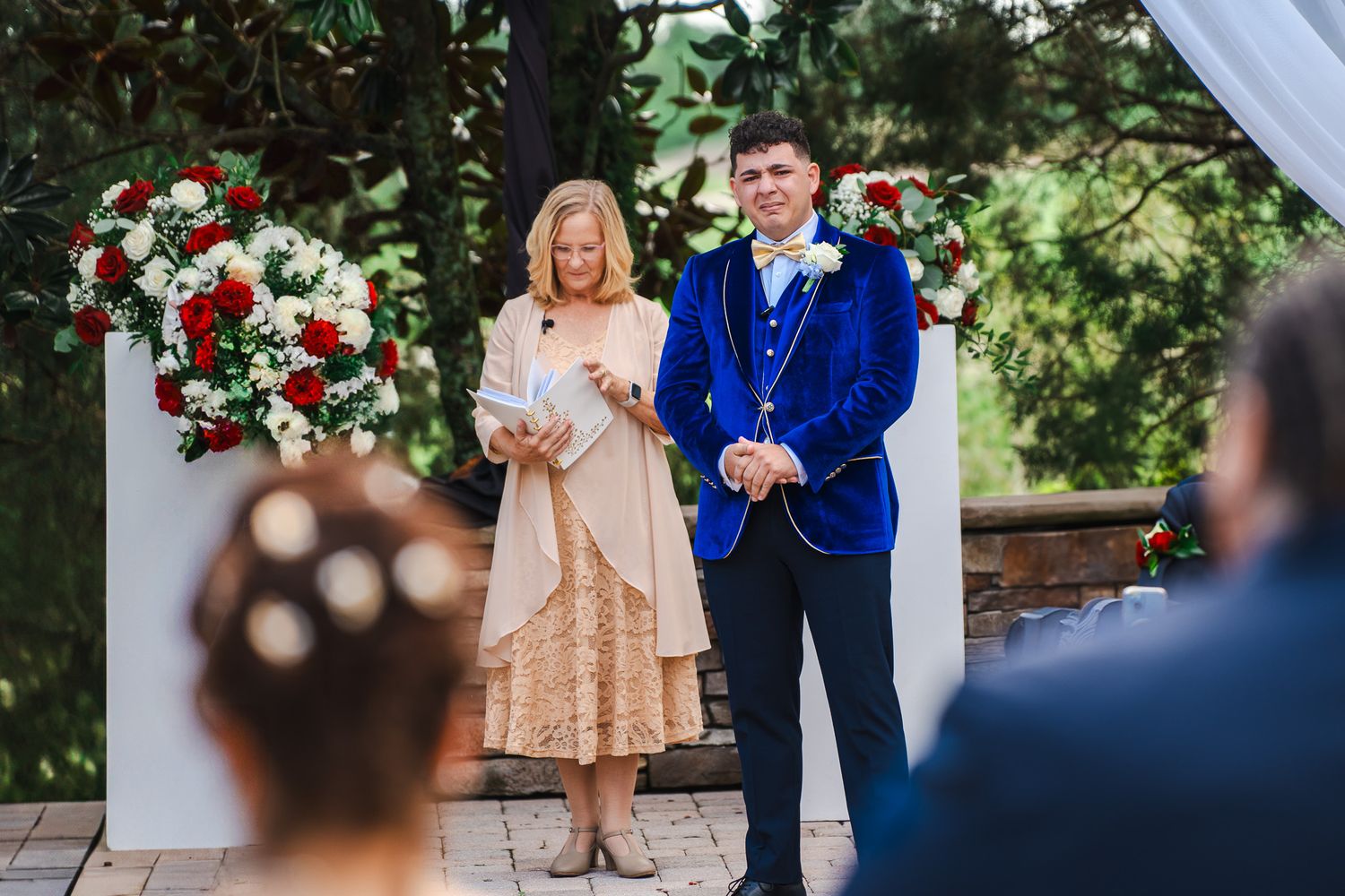 Groom in blue velvet tuxedo stands at outdoor altar with floral arrangements during ceremony.