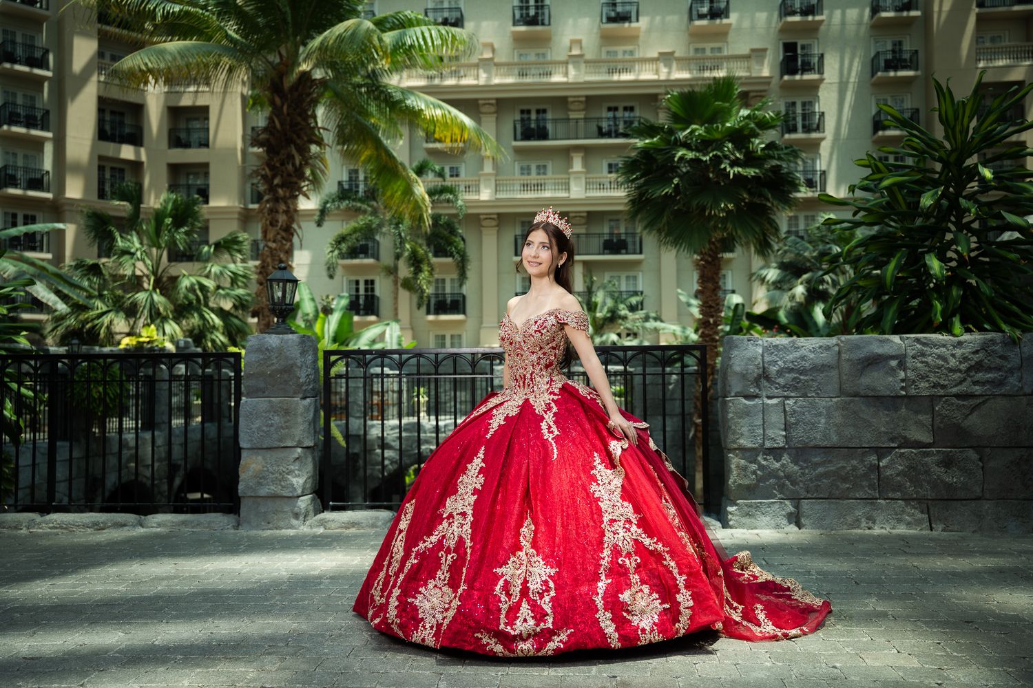 Young woman in a stunning red and gold ball gown quinceañera dress poses outdoors near tropical palms.