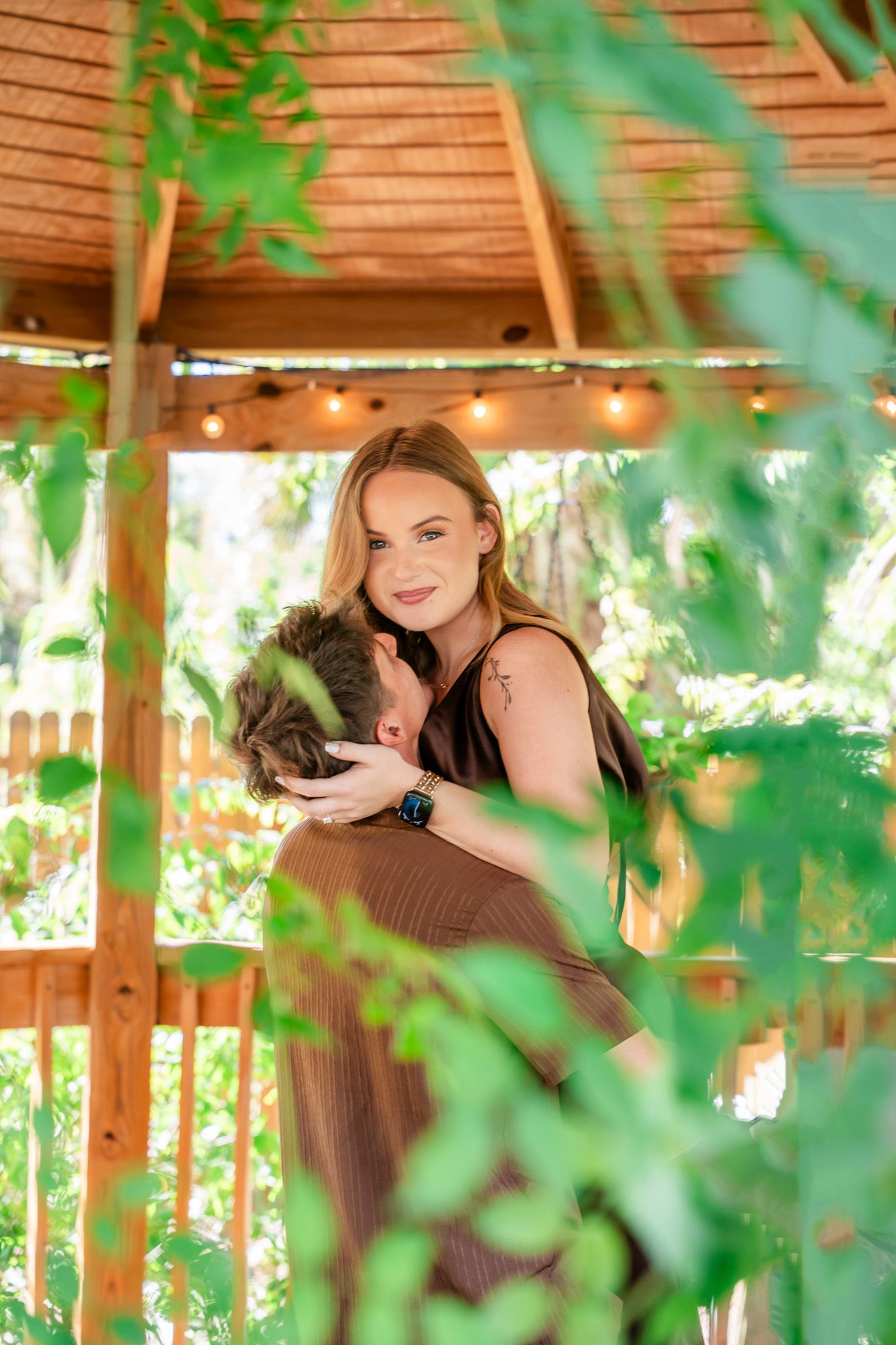 A woman in a brown dress holds a small dog under a wooden gazebo surrounded by lush green foliage.