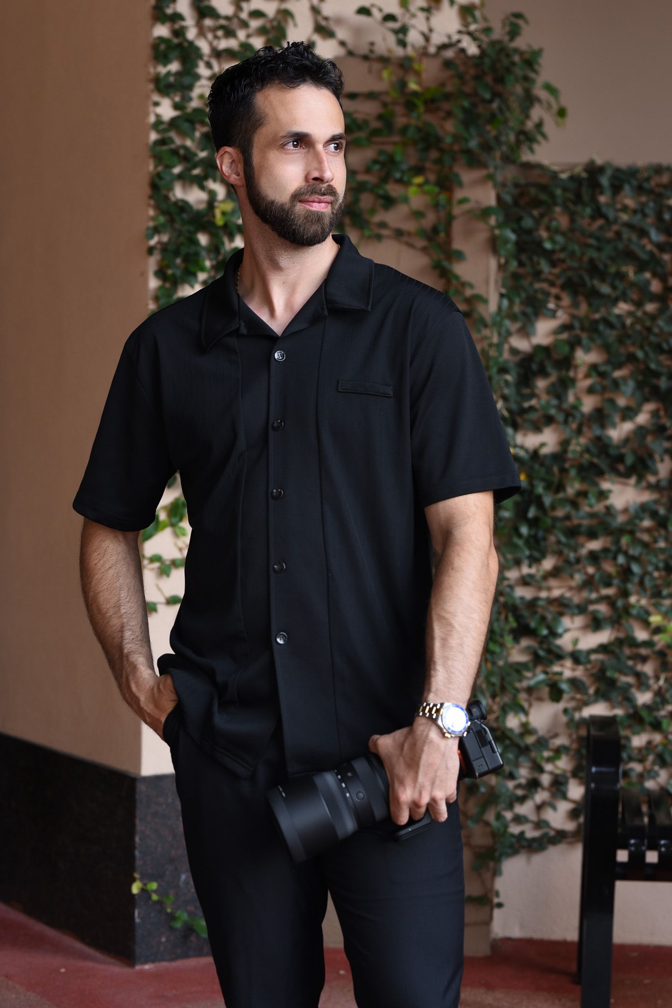 Bearded man in black shirt holding a camera outdoors near ivy-covered wall.