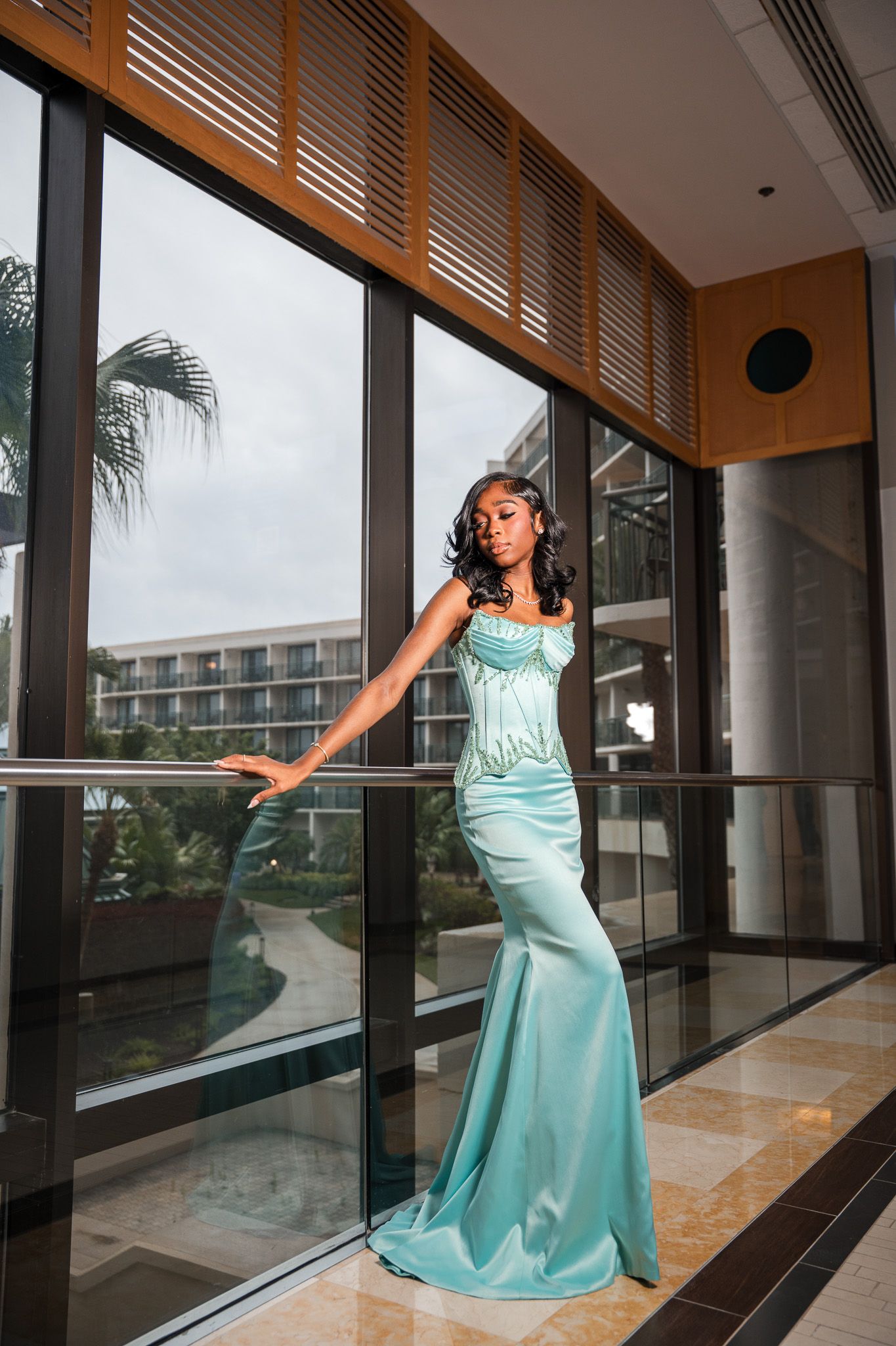 Woman in elegant teal mermaid gown posing gracefully by large windows in a modern hotel lobby.