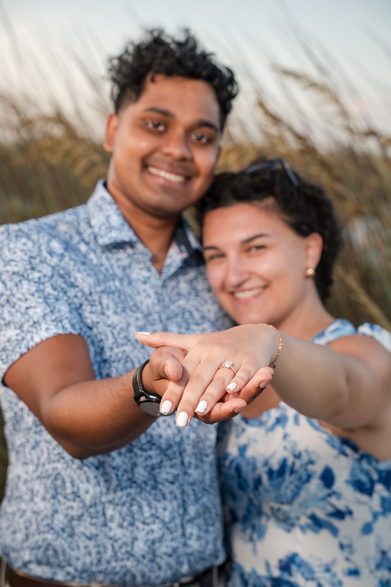 Smiling couple showing off engagement ring outdoors near tall grass at golden hour.