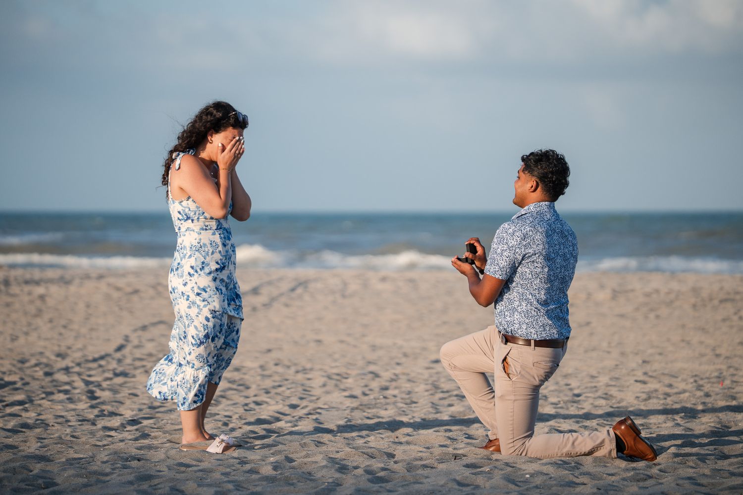Man proposing on one knee to woman on sandy beach, holding ring box as she covers her face in surprise.