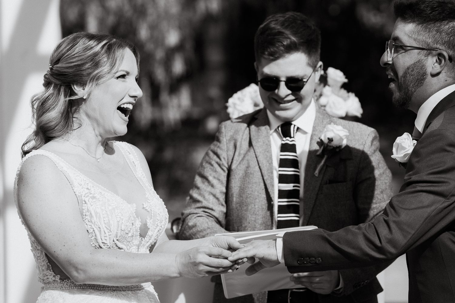 Black and white photo of bride laughing joyfully during wedding ring exchange ceremony with officiant.