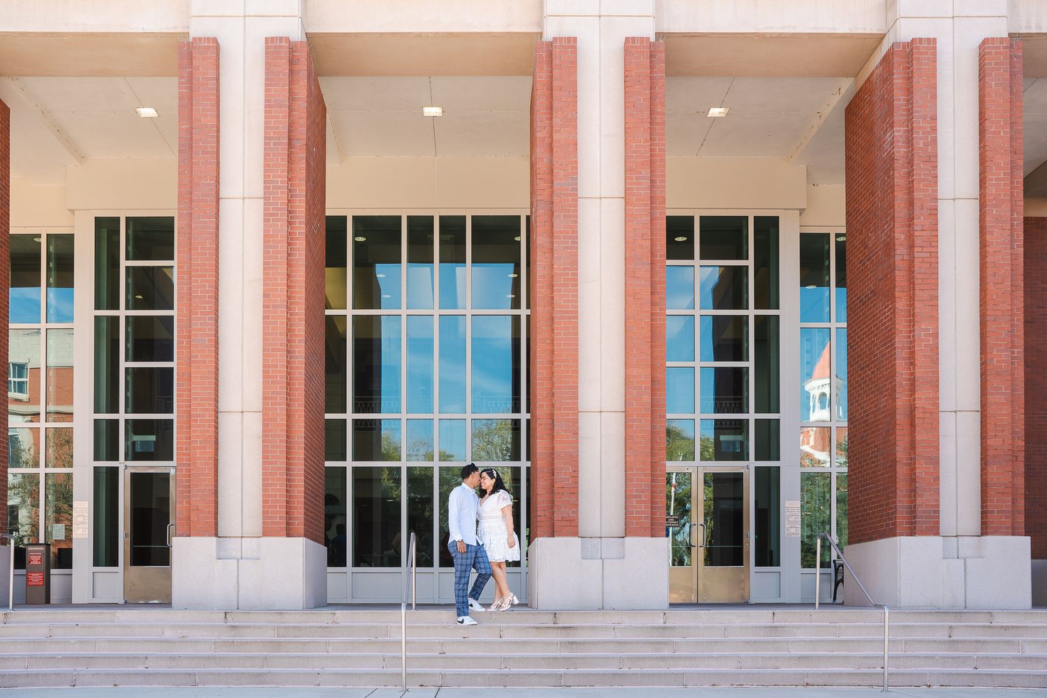 Couple shares a kiss on the steps of a grand brick building with tall glass windows and columns.