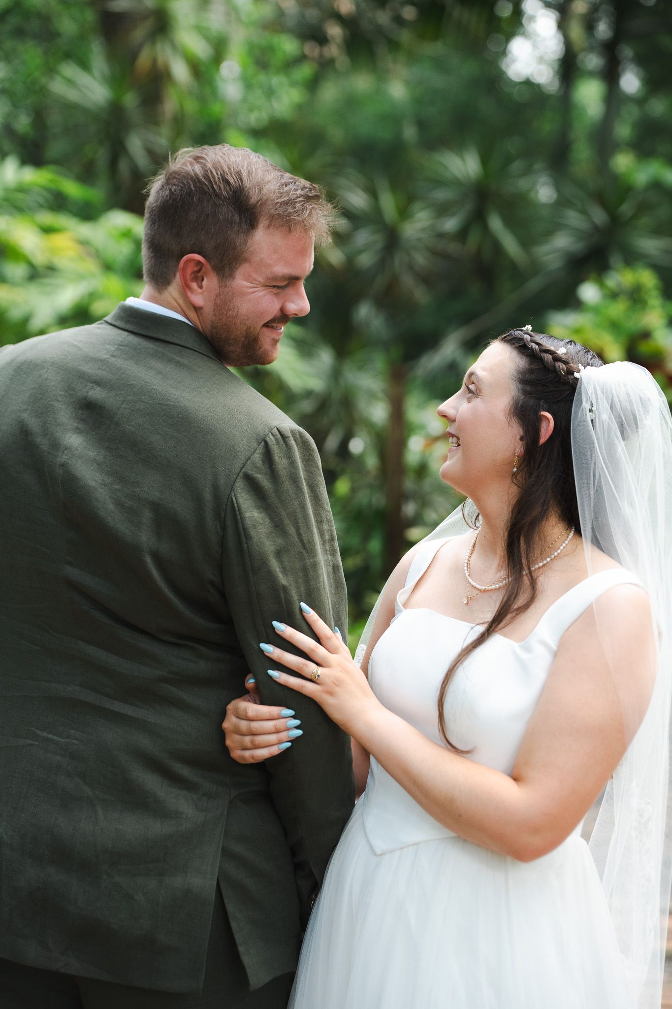 Bride in white gown smiles at groom in olive suit during an intimate outdoor garden wedding portrait.