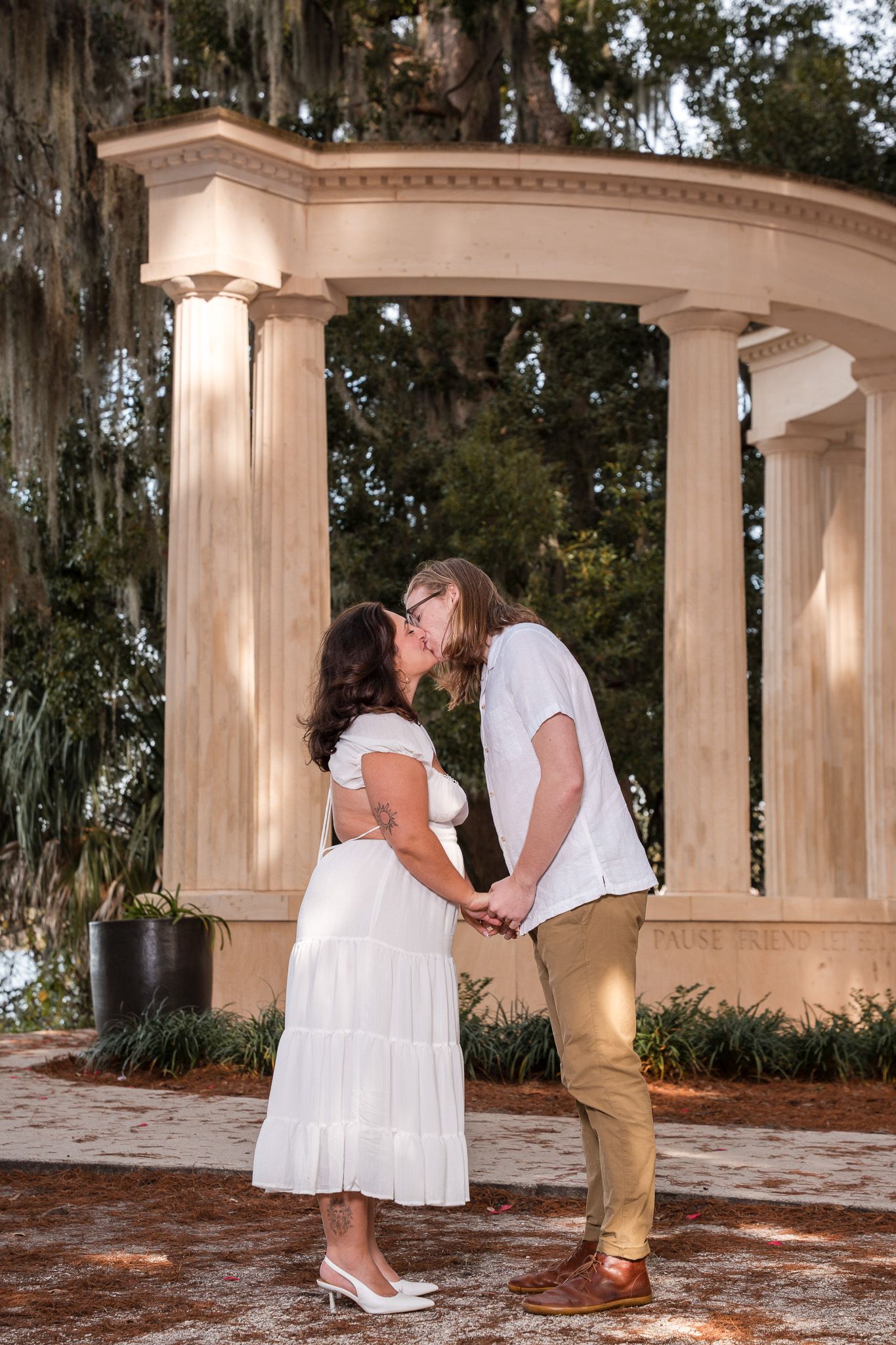 Couple sharing a kiss in front of elegant stone columns with Spanish moss-covered trees in background.