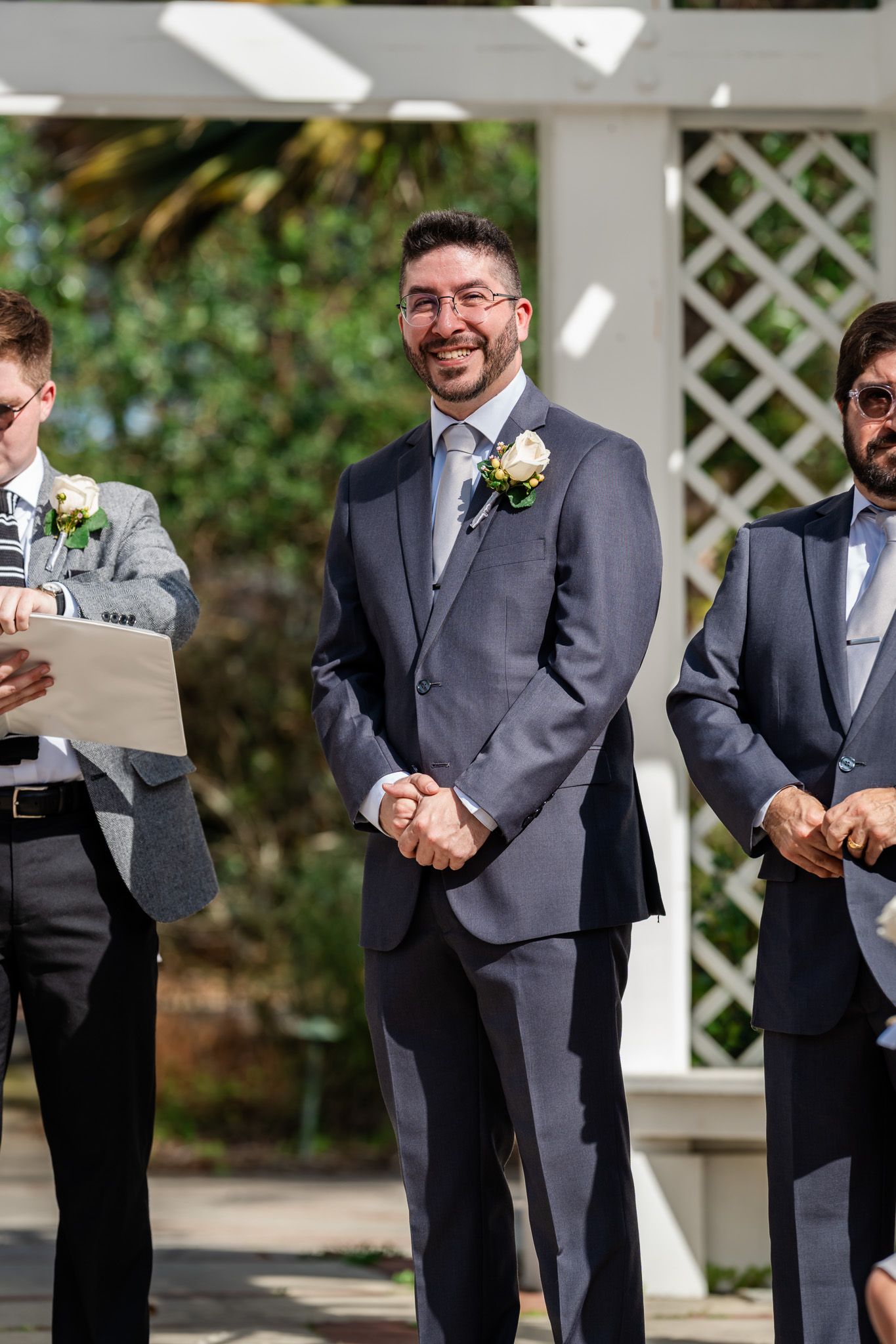 Smiling groom in a gray suit with boutonniere stands at the altar with groomsmen at an outdoor wedding.