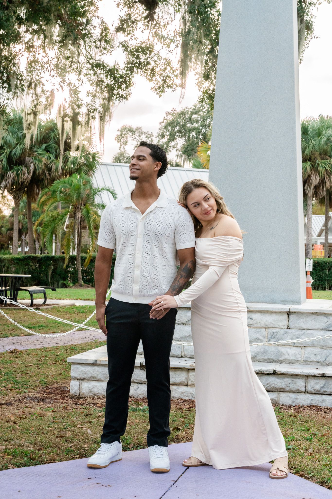 Interracial couple posing by a column in a park, woman in white gown leaning on mans shoulder.