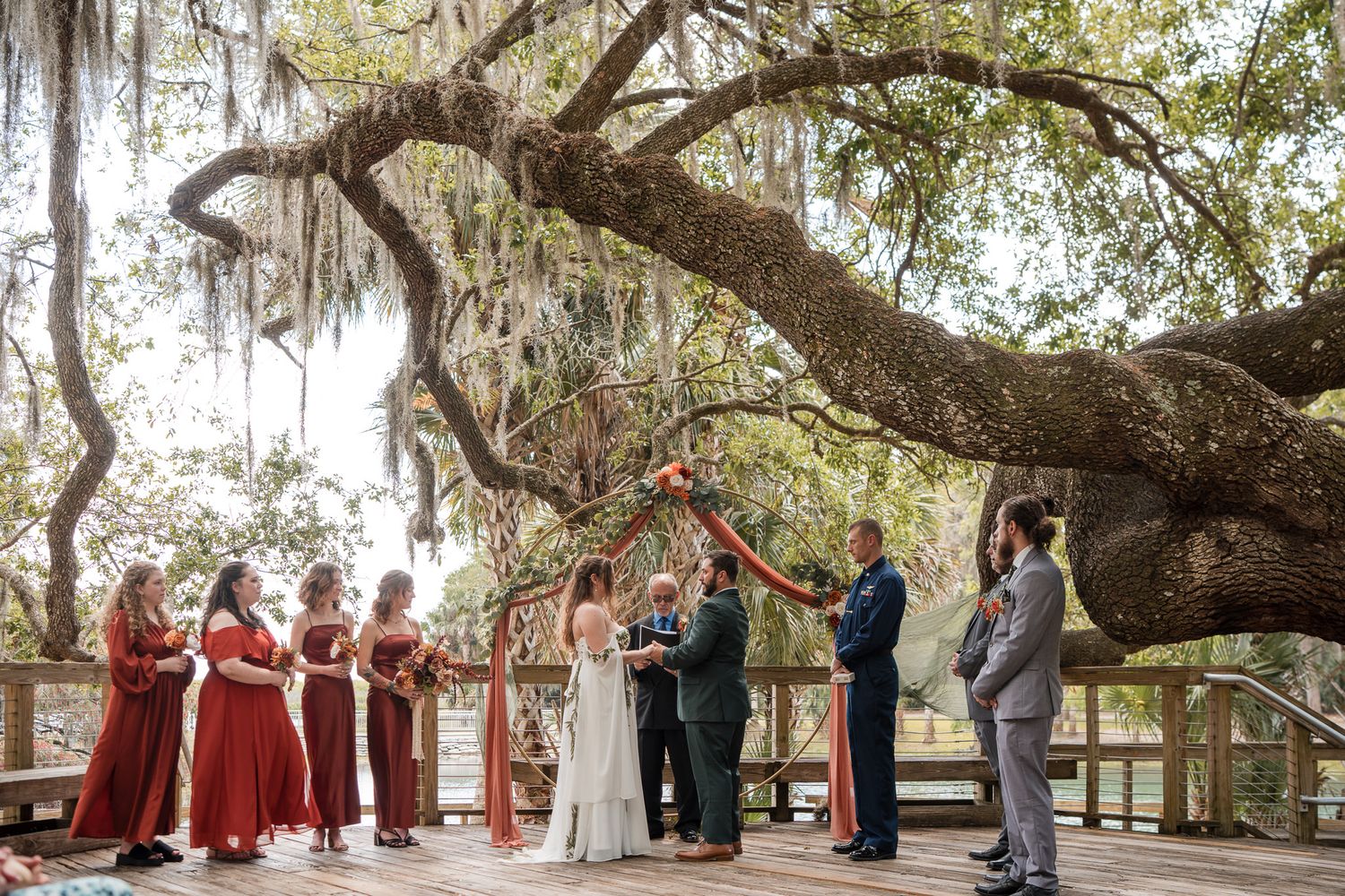 Outdoor wedding ceremony on a wooden deck under a large oak tree with Spanish moss, featuring bridesmaids in red dresses.