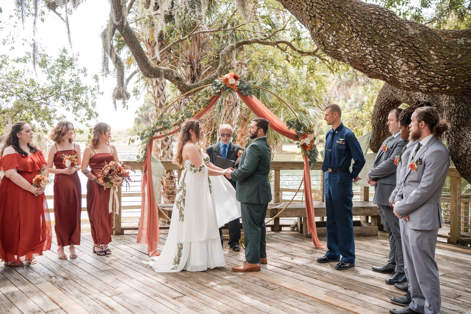 Outdoor wedding ceremony on wooden deck under oak tree with bridesmaids in red dresses and groomsmen in suits.