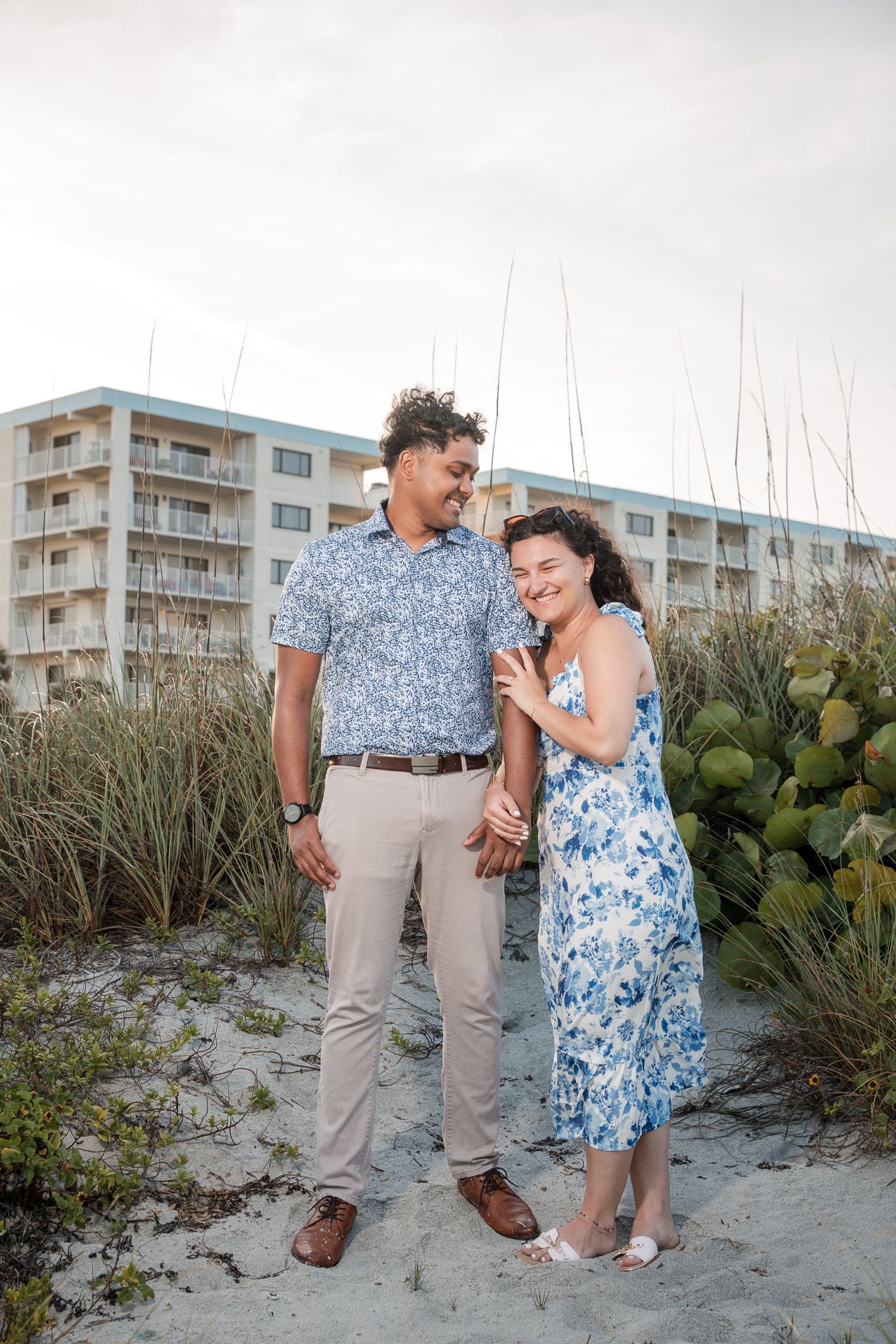 A couple holds hands and smiles on a sandy beach, dressed in matching blue floral outfits with a hotel in the background.