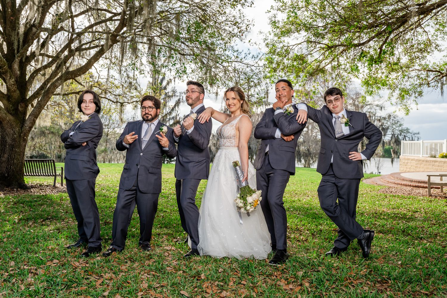 Bride with bouquet poses playfully with five groomsmen in gray suits striking fun poses under a large oak tree.