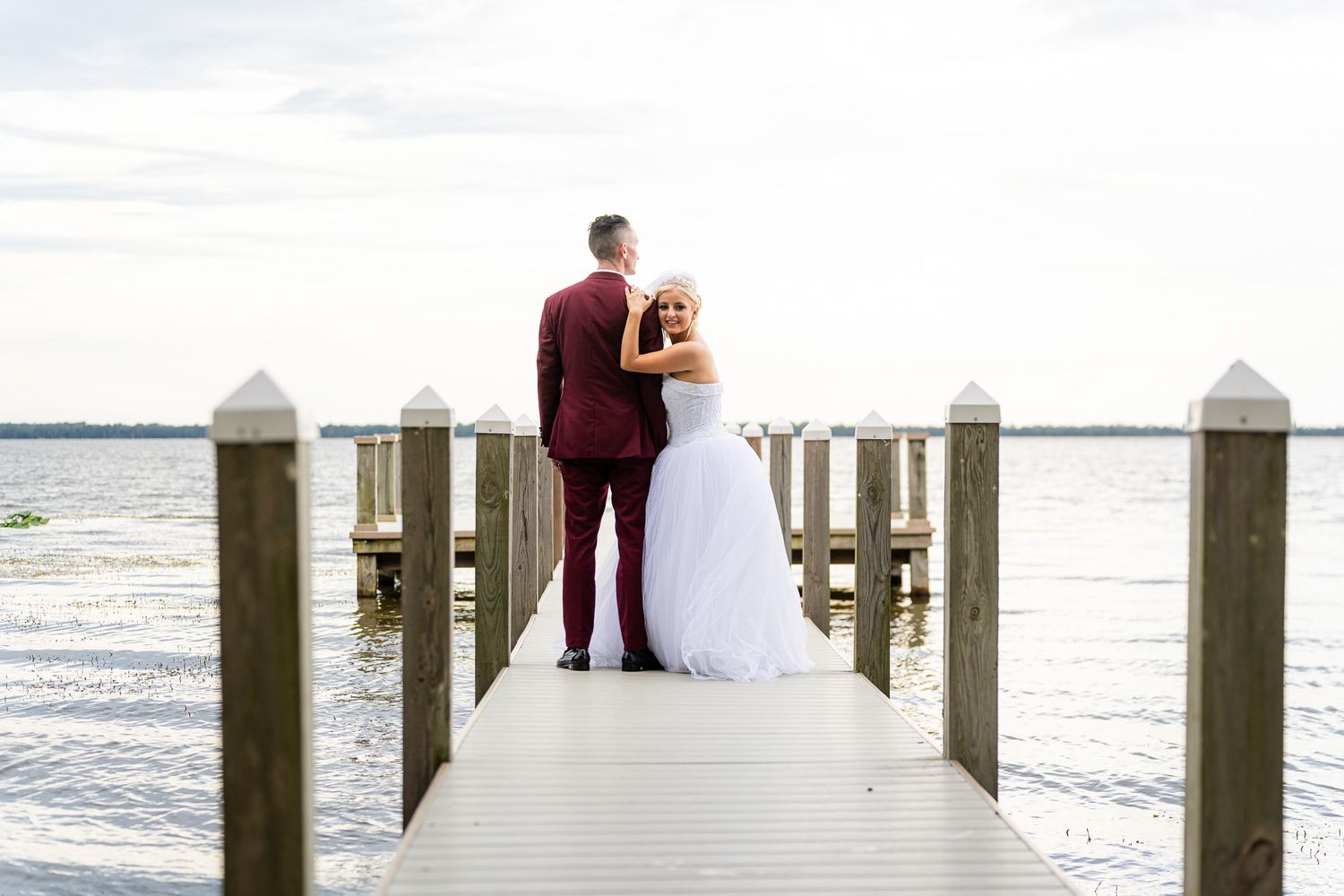 Bride and groom standing together on wooden dock overlooking calm lake at sunset.