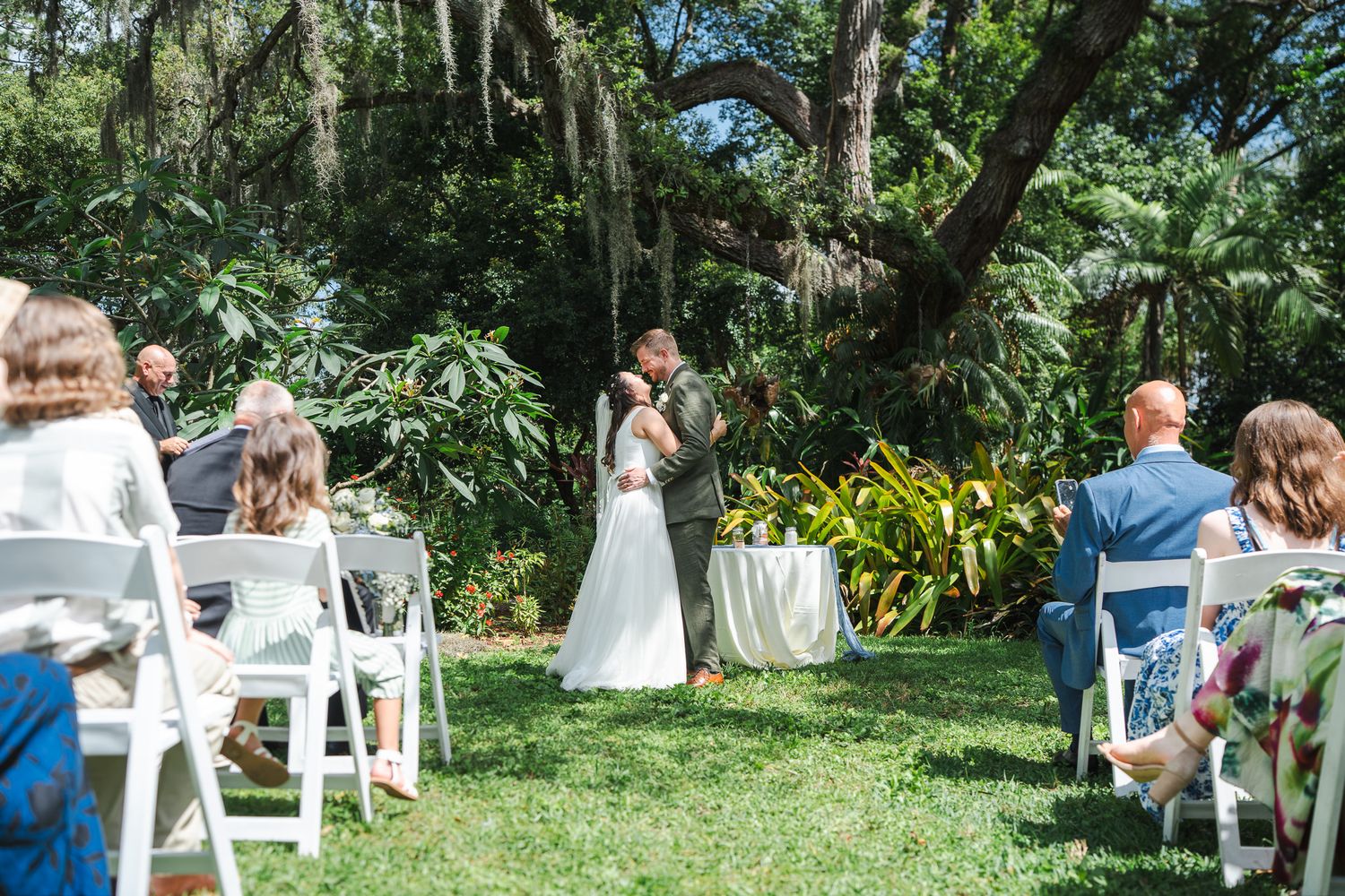 Outdoor garden wedding ceremony with bride and groom at the altar surrounded by lush tropical greenery and seated guests.