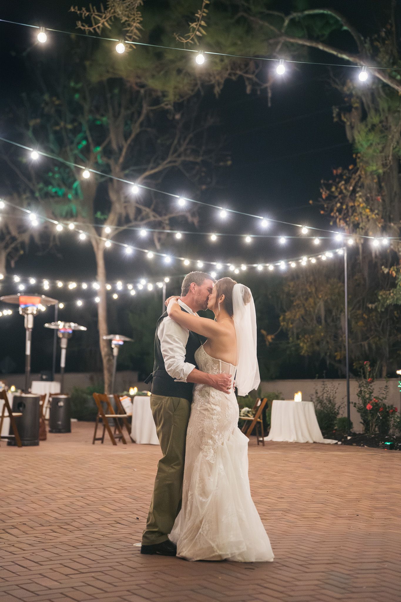 Newlyweds share romantic first dance under glowing string lights at outdoor night reception.