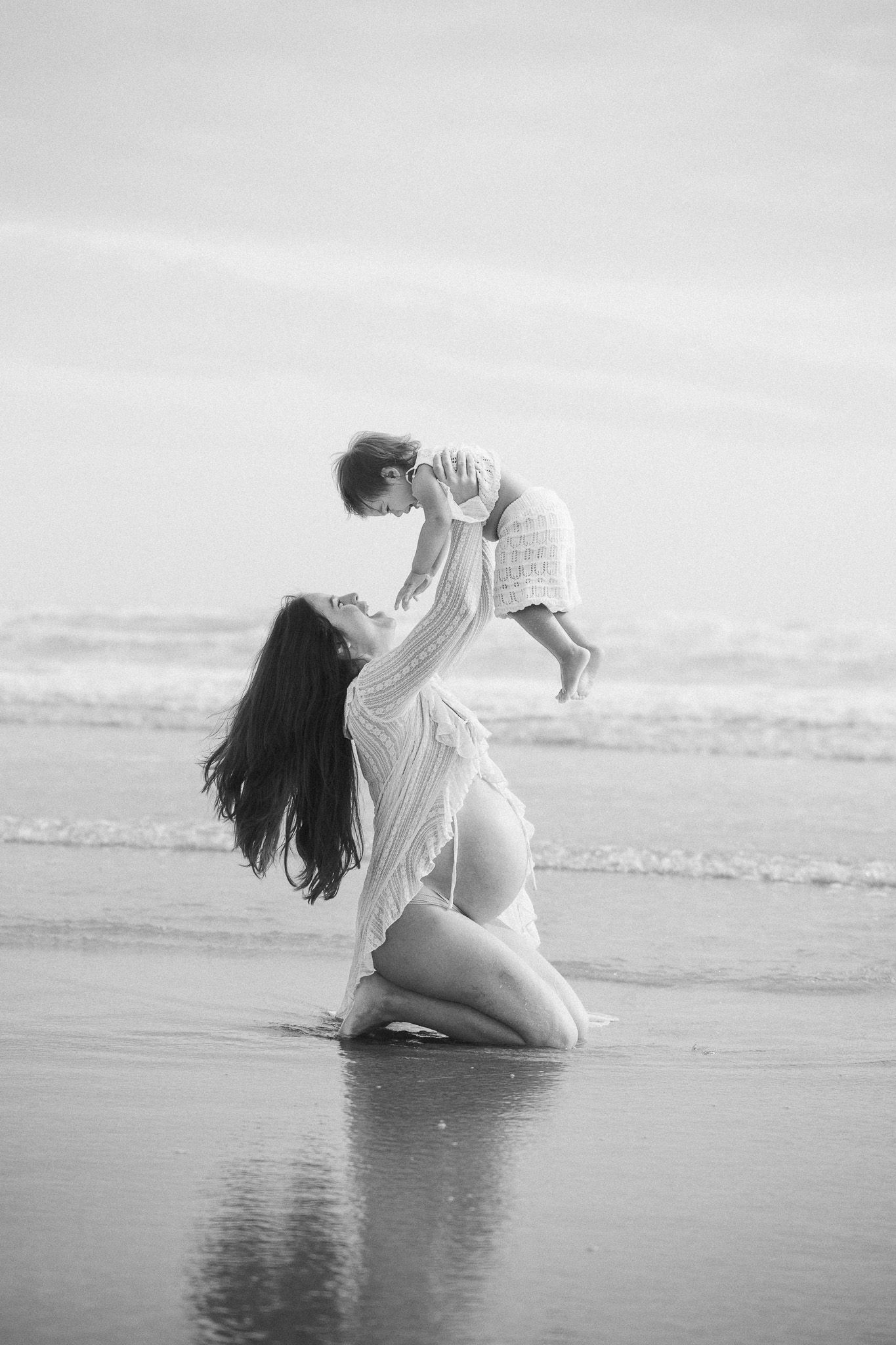 Pregnant mother kneeling on beach lifting baby in black and white maternity photo.