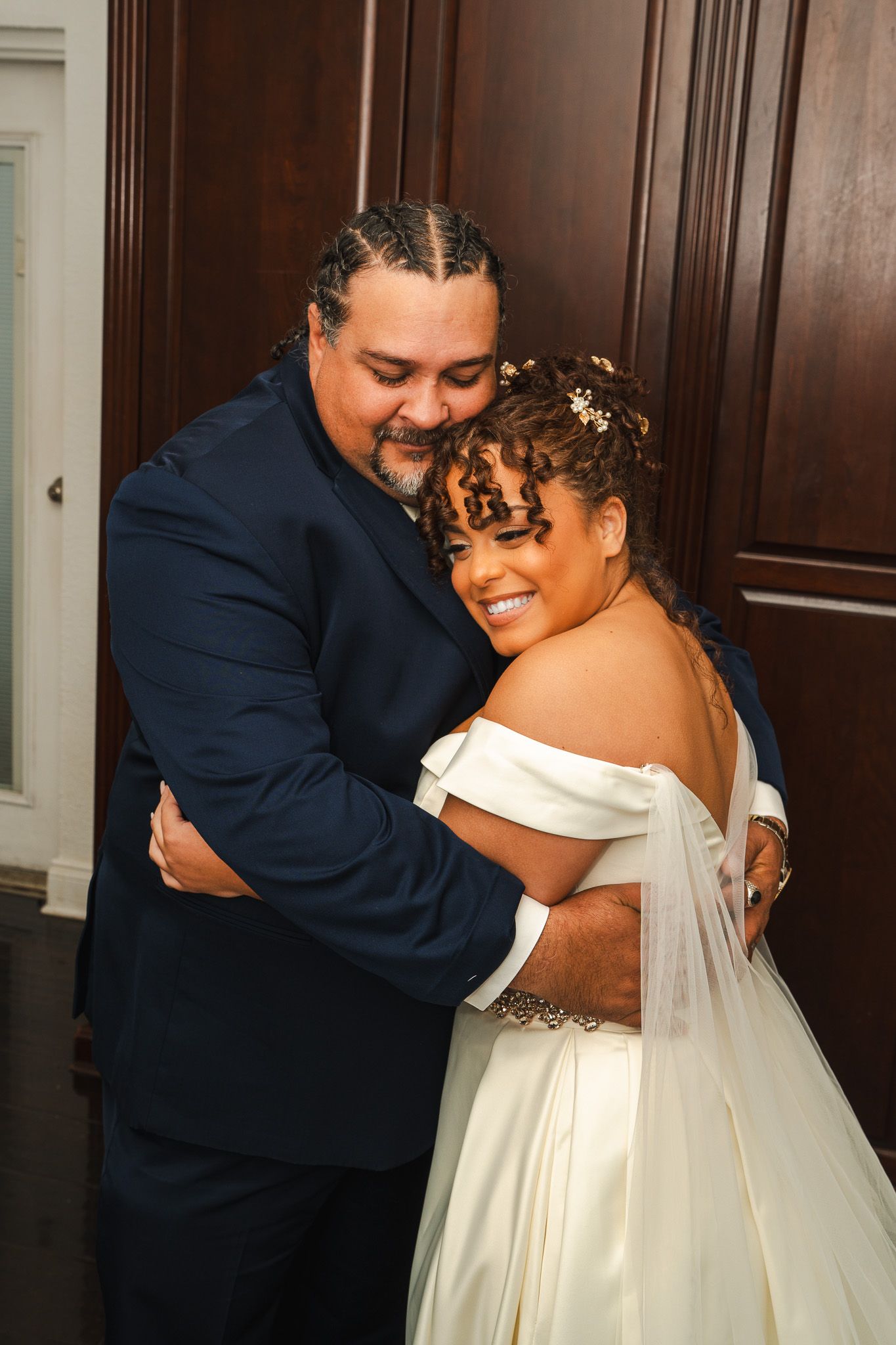 Emotional father embraces smiling bride in off-shoulder wedding gown before ceremony.