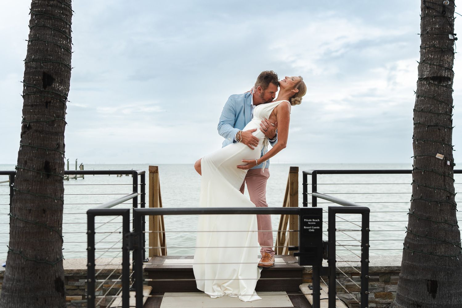 Groom dips bride for romantic kiss between two palm trees on wooden dock overlooking the ocean.