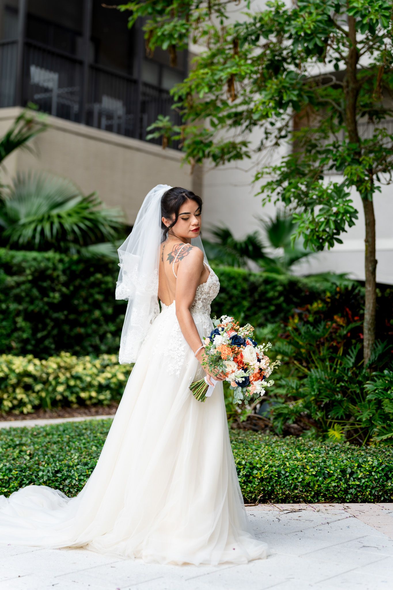 Bride in backless lace wedding dress with veil and tattoo holds bouquet in lush garden setting.