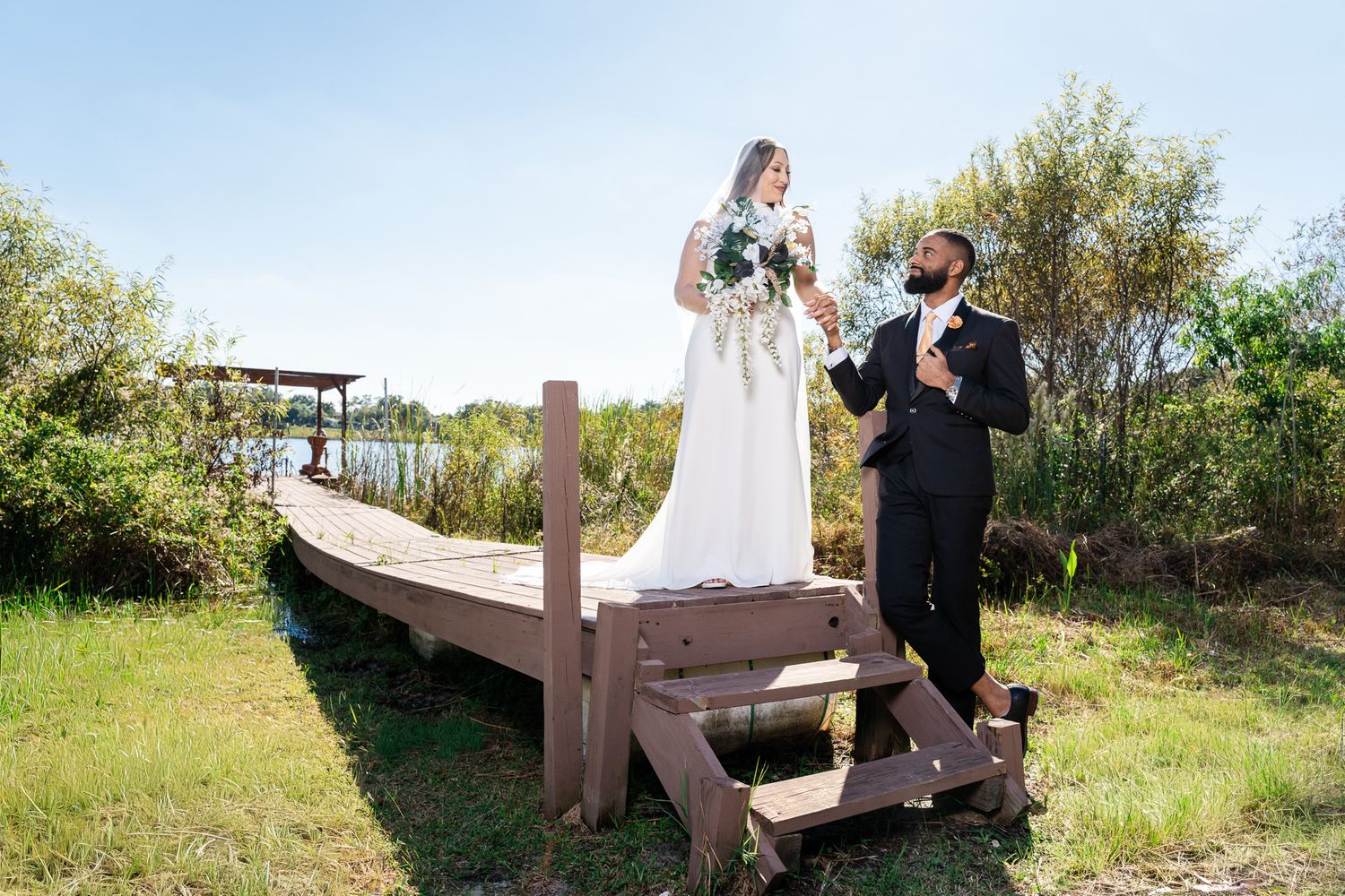 Bride stands elevated on wooden structure holding bouquet while groom adjusts tie by a lakeside.