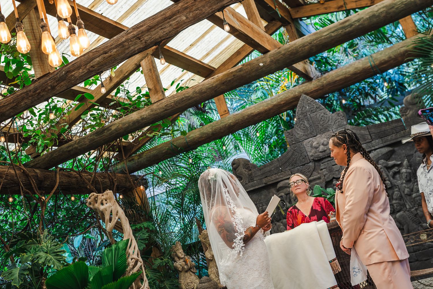 Wide shot of same-sex wedding ceremony under rustic wooden pergola with tropical garden backdrop.