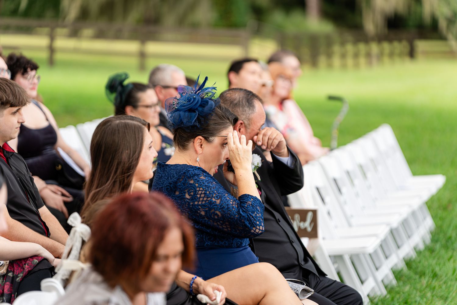Emotional wedding guest in navy lace dress wipes tears during outdoor ceremony.