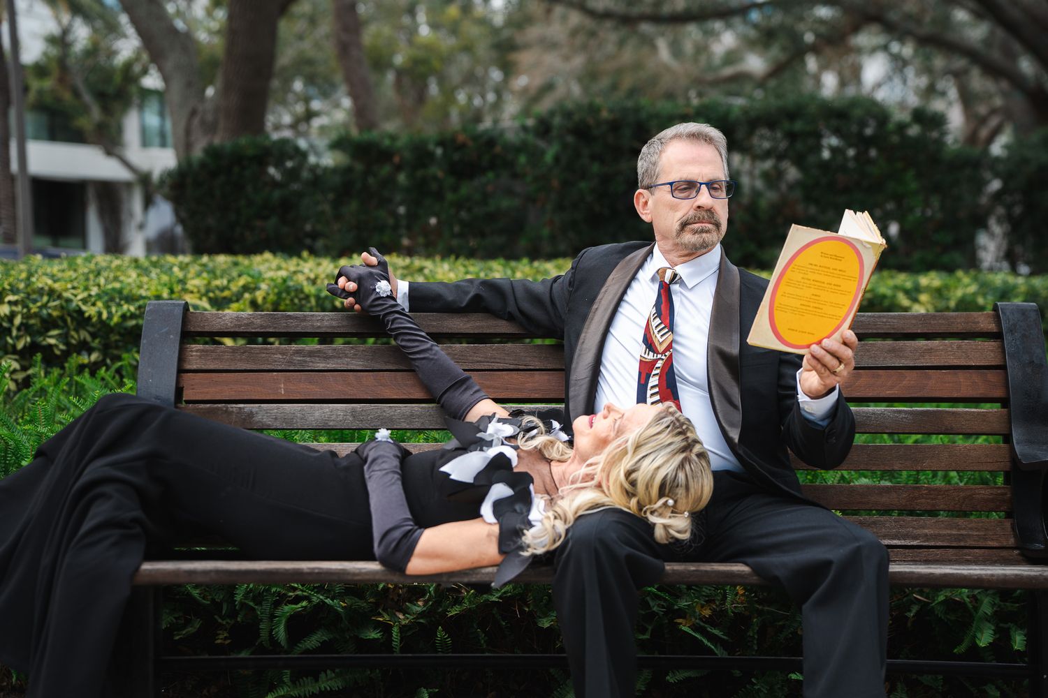 Couple poses humorously on a park bench, woman lying across mans lap as he reads a yellow book in a suit.