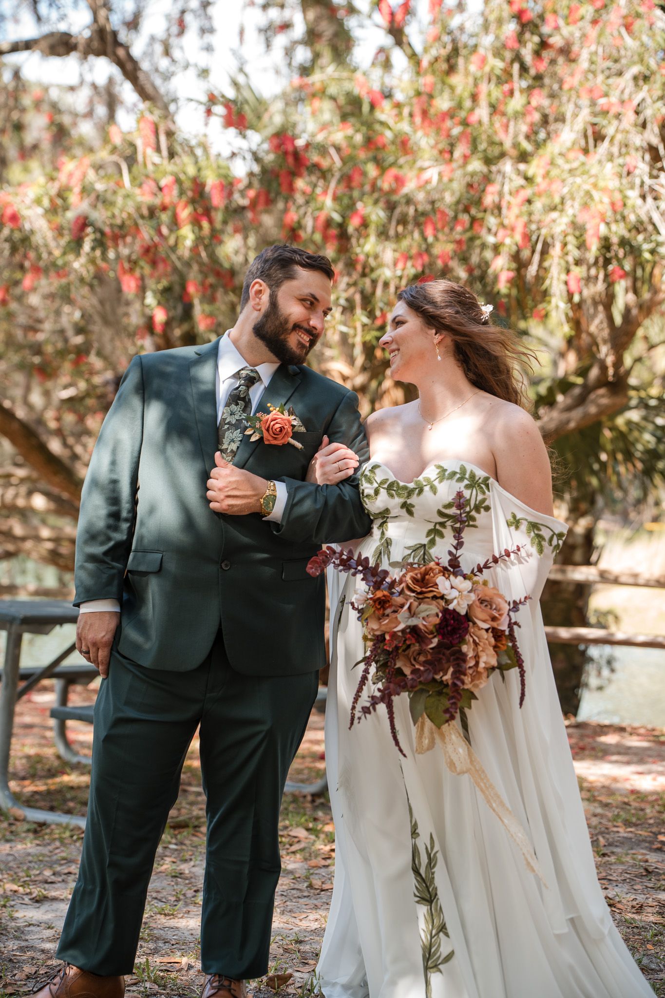 A bride and groom share a joyful moment outdoors, she holds a bouquet, he wears a green suit, with red flowers behind them.