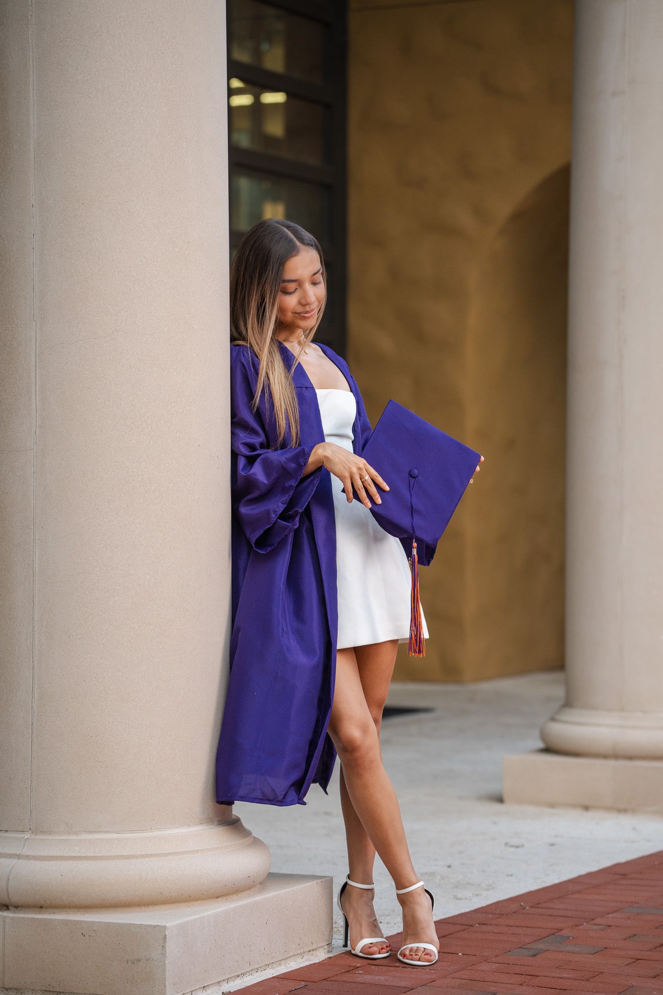 Female graduate in purple gown leans against column holding cap while looking down thoughtfully.