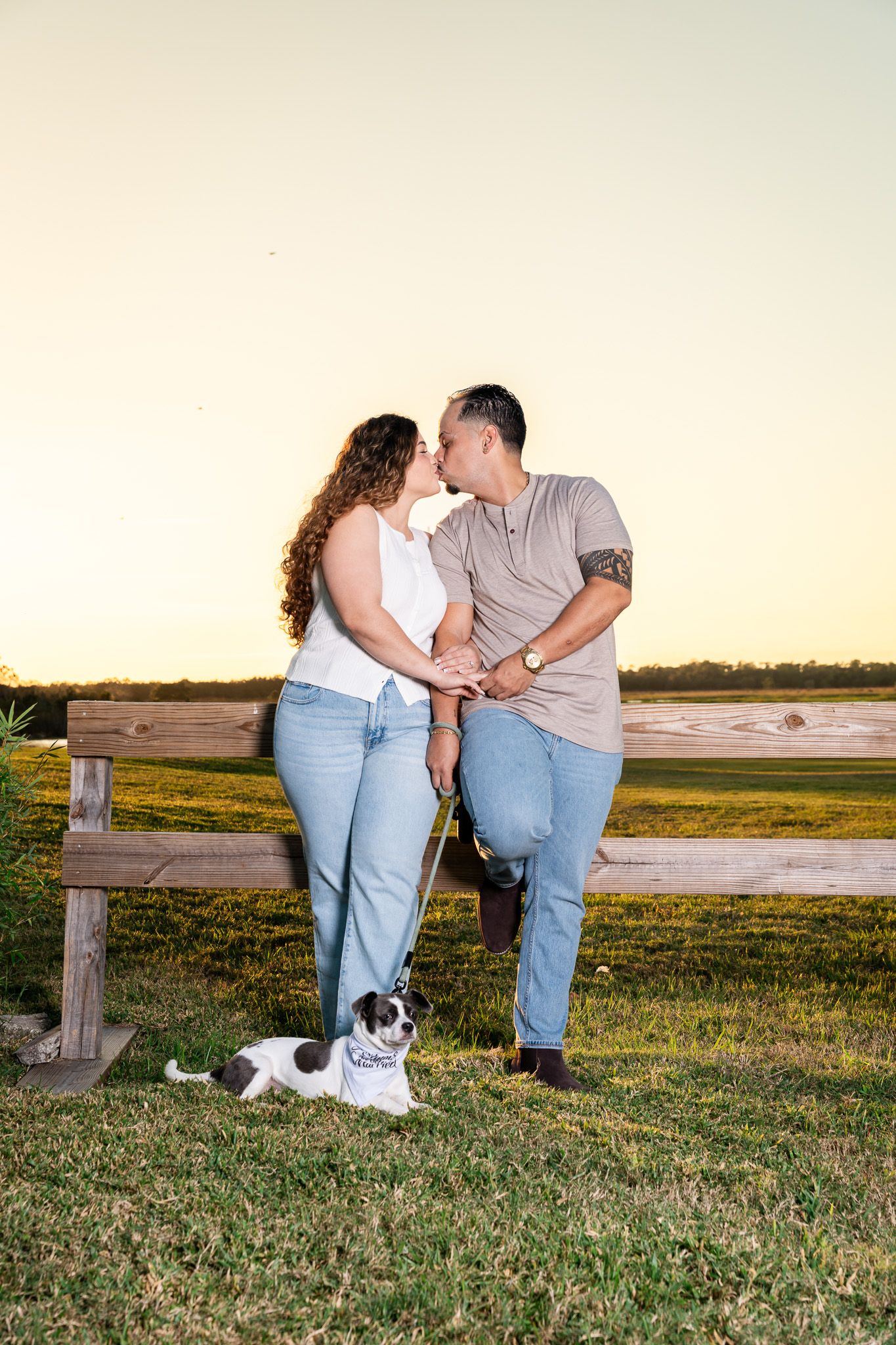 Couple sharing a kiss near a wooden fence at golden hour with their small dog on a leash.