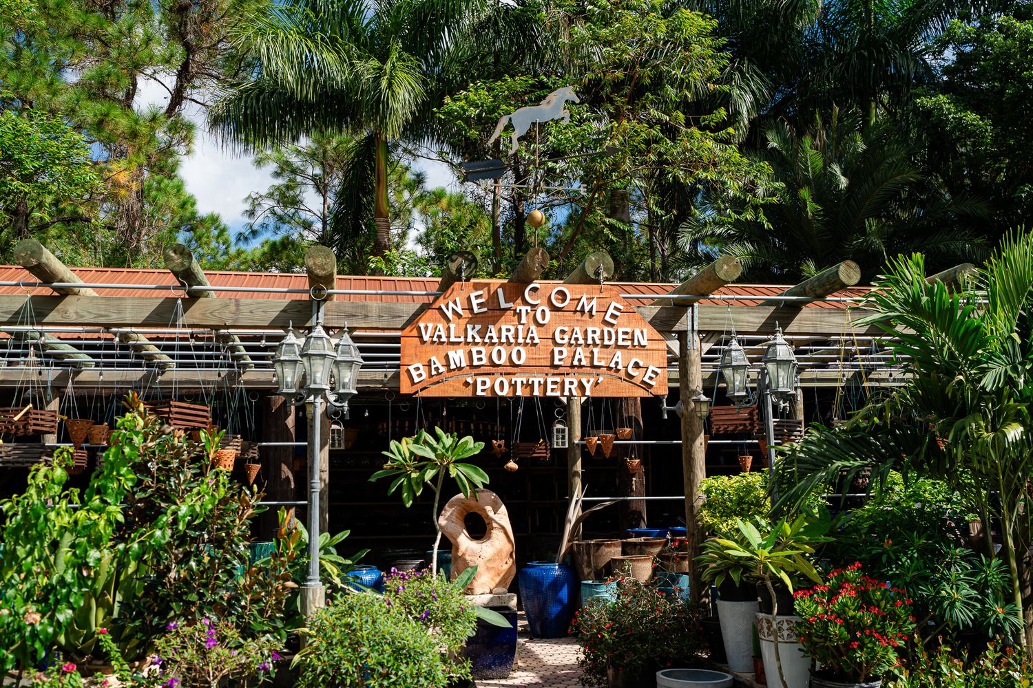 Rustic entrance sign welcoming visitors to Vilkara Gardens Bamboo Palace Pottery surrounded by lush plants.