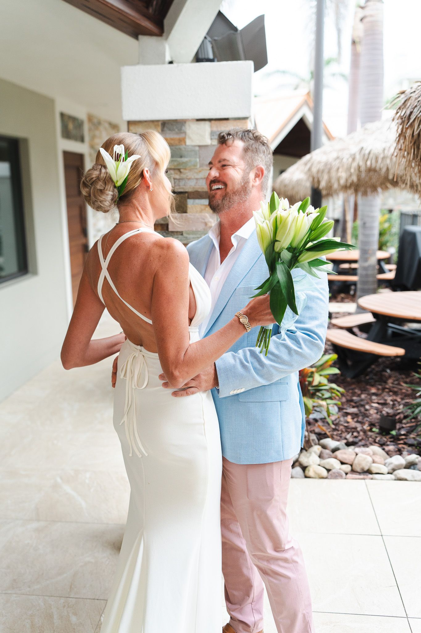 Smiling groom in light blue blazer presents white lily bouquet to bride in backless wedding dress.