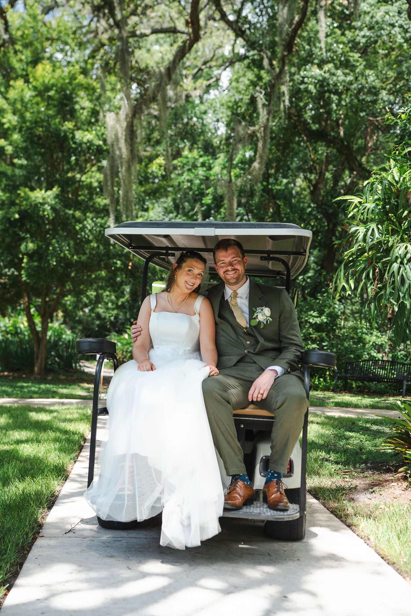 Bride and groom smile while seated together on a golf cart surrounded by lush green trees.