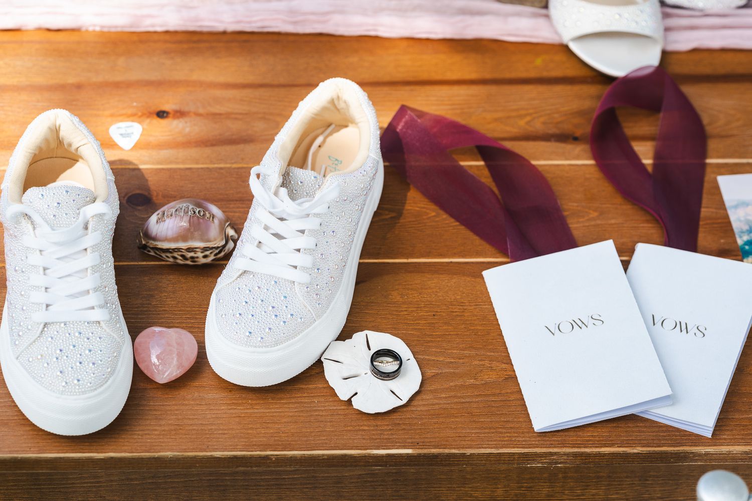 Bridal flat lay featuring white sneakers, vow books, wedding rings, and burgundy ribbon on wood.