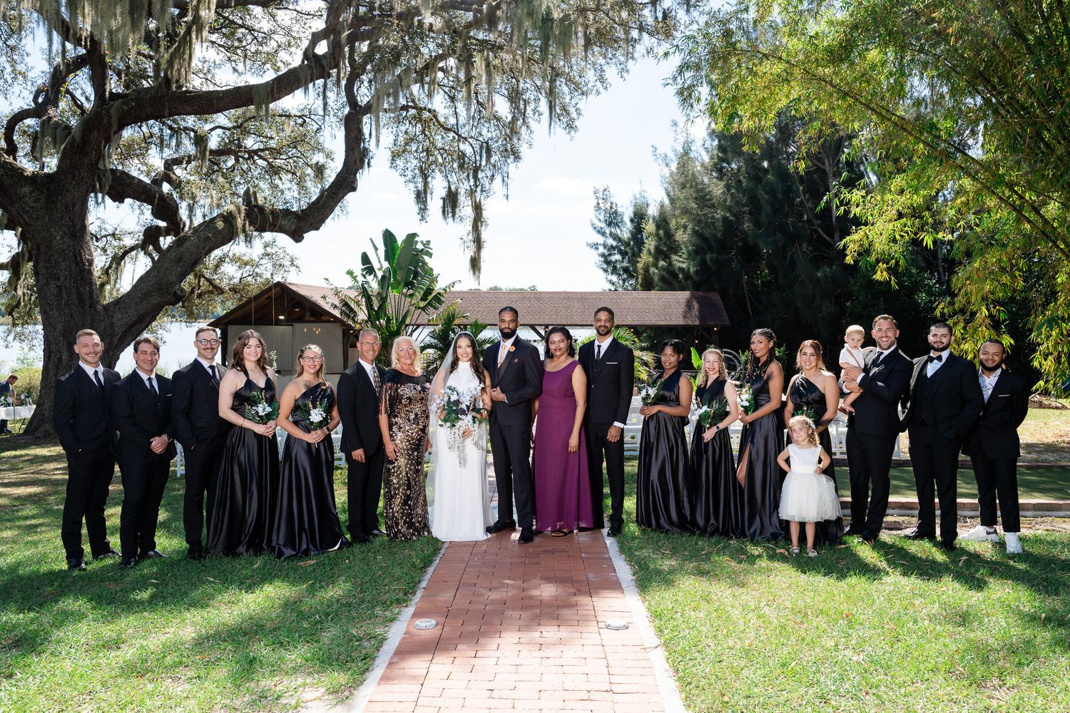 Large wedding party in black formal wear poses with bride and groom on brick path under oak trees.