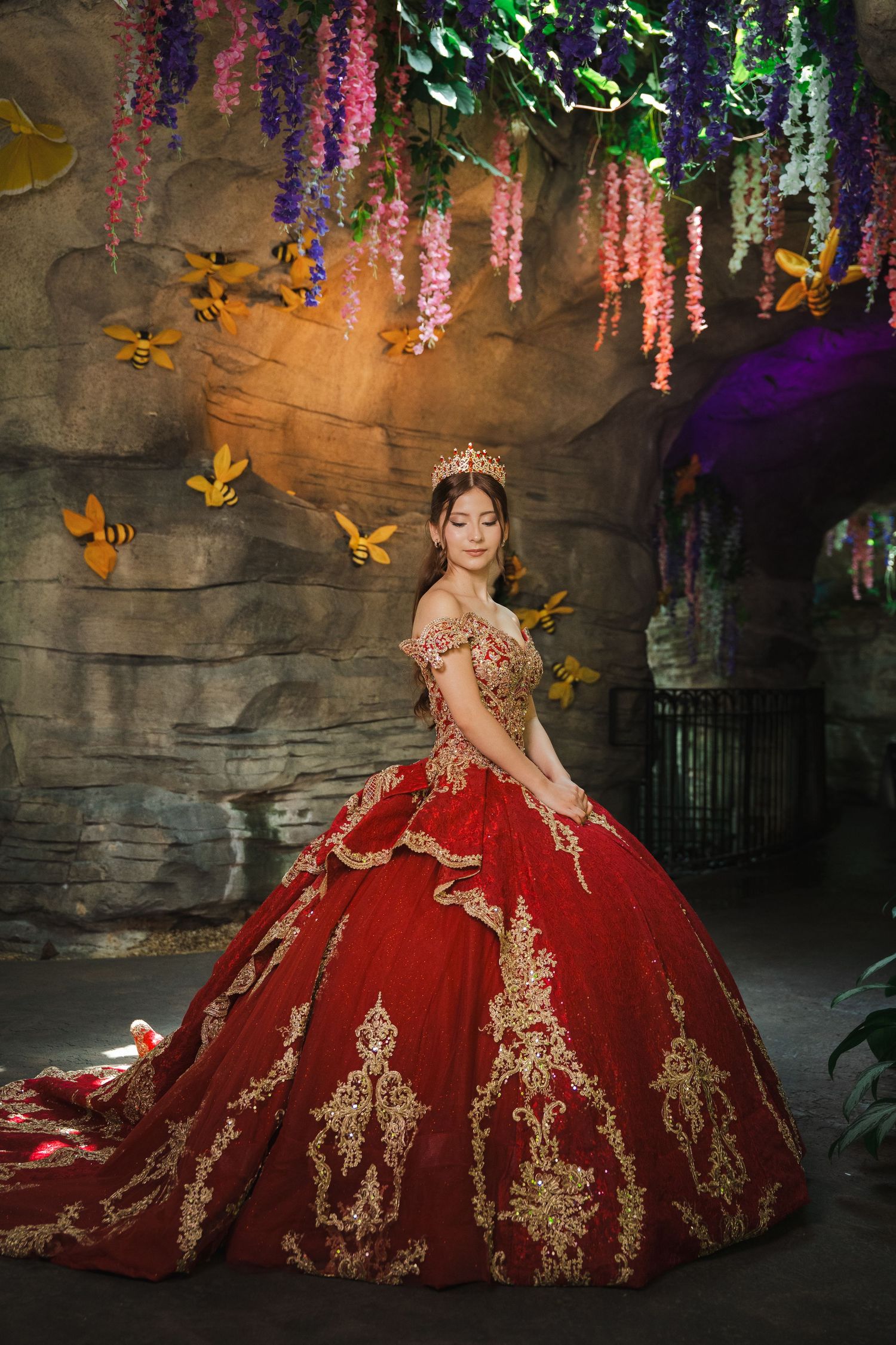 Young woman in red gold-embroidered ball gown and crown poses in enchanted garden setting with hanging flowers.