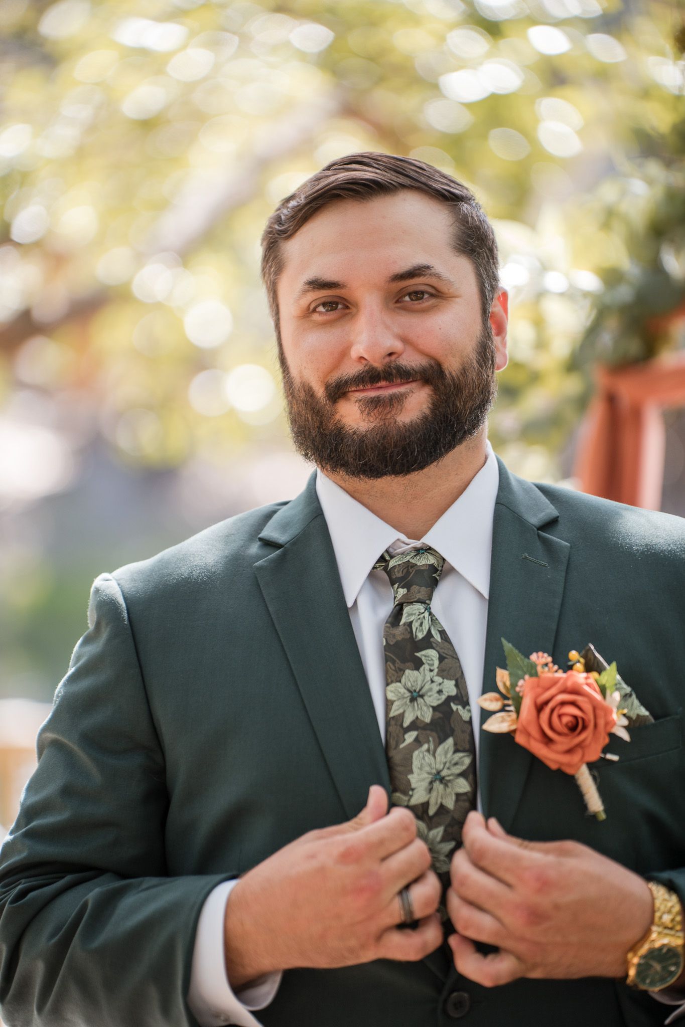 Bearded groom in green suit adjusting floral tie with orange rose boutonniere at outdoor wedding.
